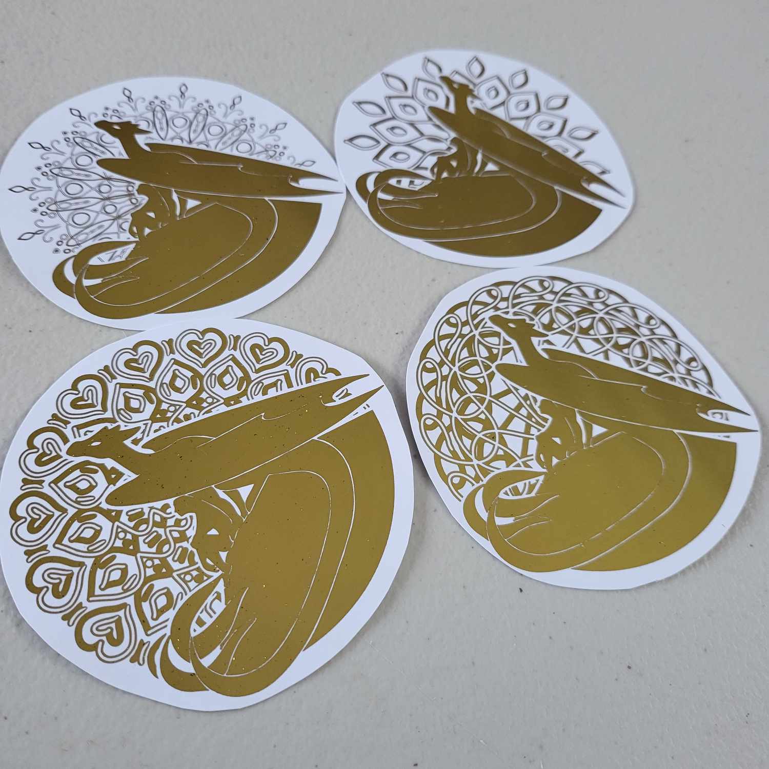 Four round coaster-sized gold foiled dragons on paper. The dragons are all identical, but they have different background designs.