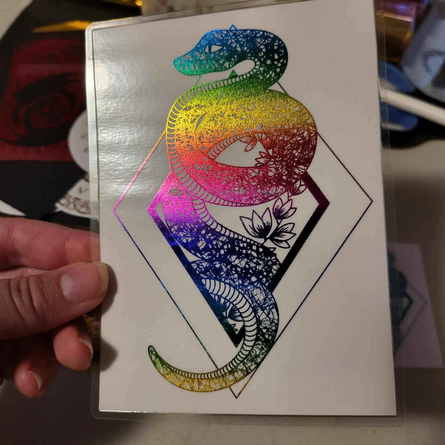 A close-up of the laminated rainbow-foiled snake design on white card. It is held so that the light is reflecting off the metallic foil.