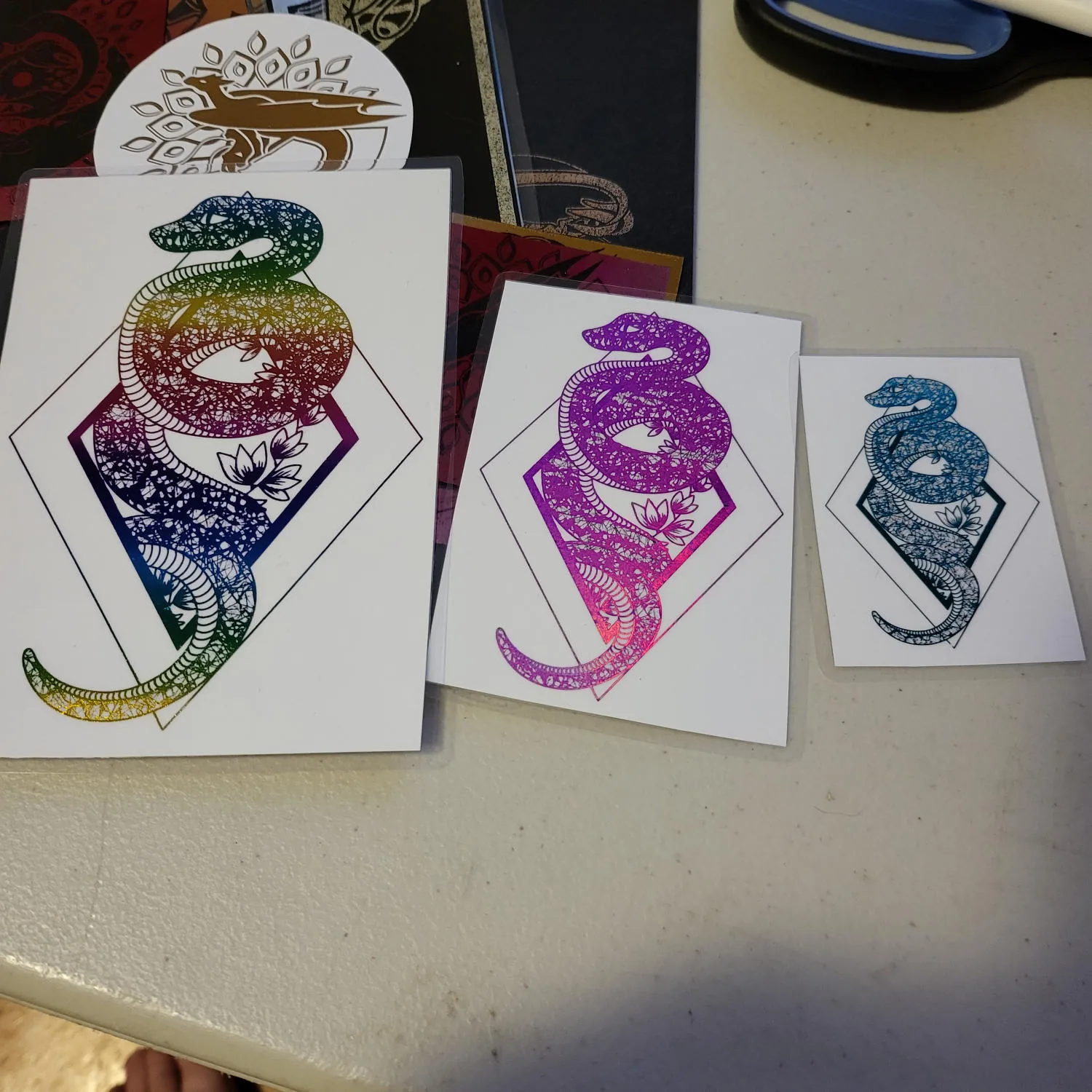 Three laminated foiled snake designs on white card of different sizes. The largest is foiled in rainbow gradient, the medium one has pink foil, the smallest has dark blue foil.