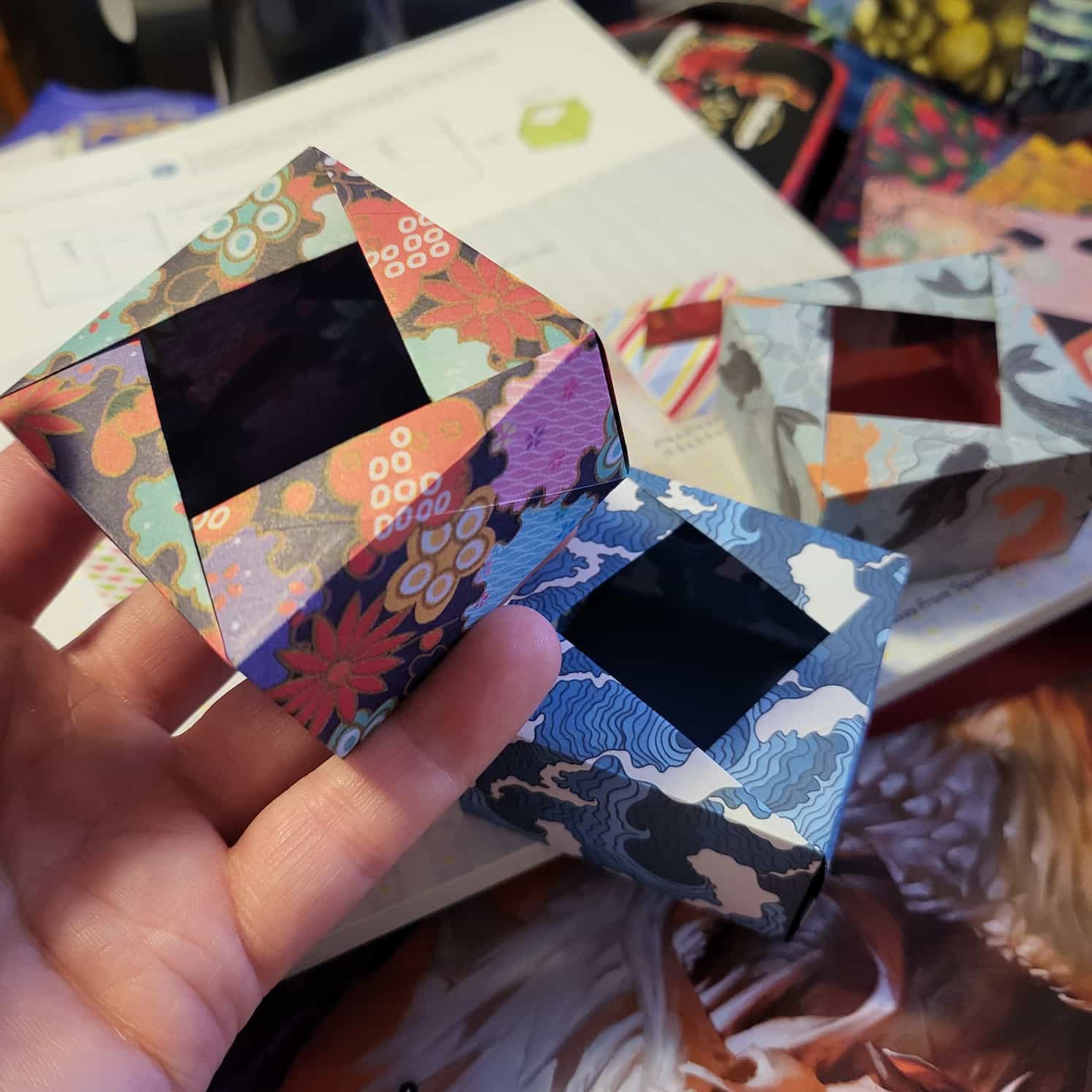Three completed origami boxes with offset diagonal openings. In the background is the koi box, and one with a blue and white waves design. A hand is holding one box closer made with a colourful floral design.