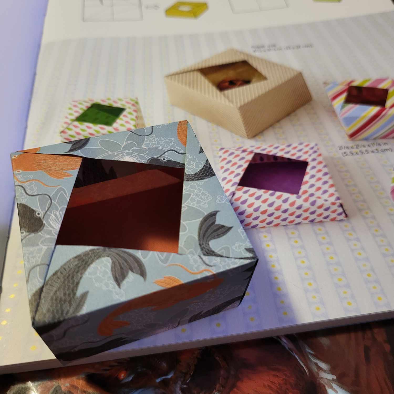 An origami box with offset diagonal opening. The paper is blue with black and orange koi. The inside of the box is orange. In the background you see the book with instructions and images of other boxes.