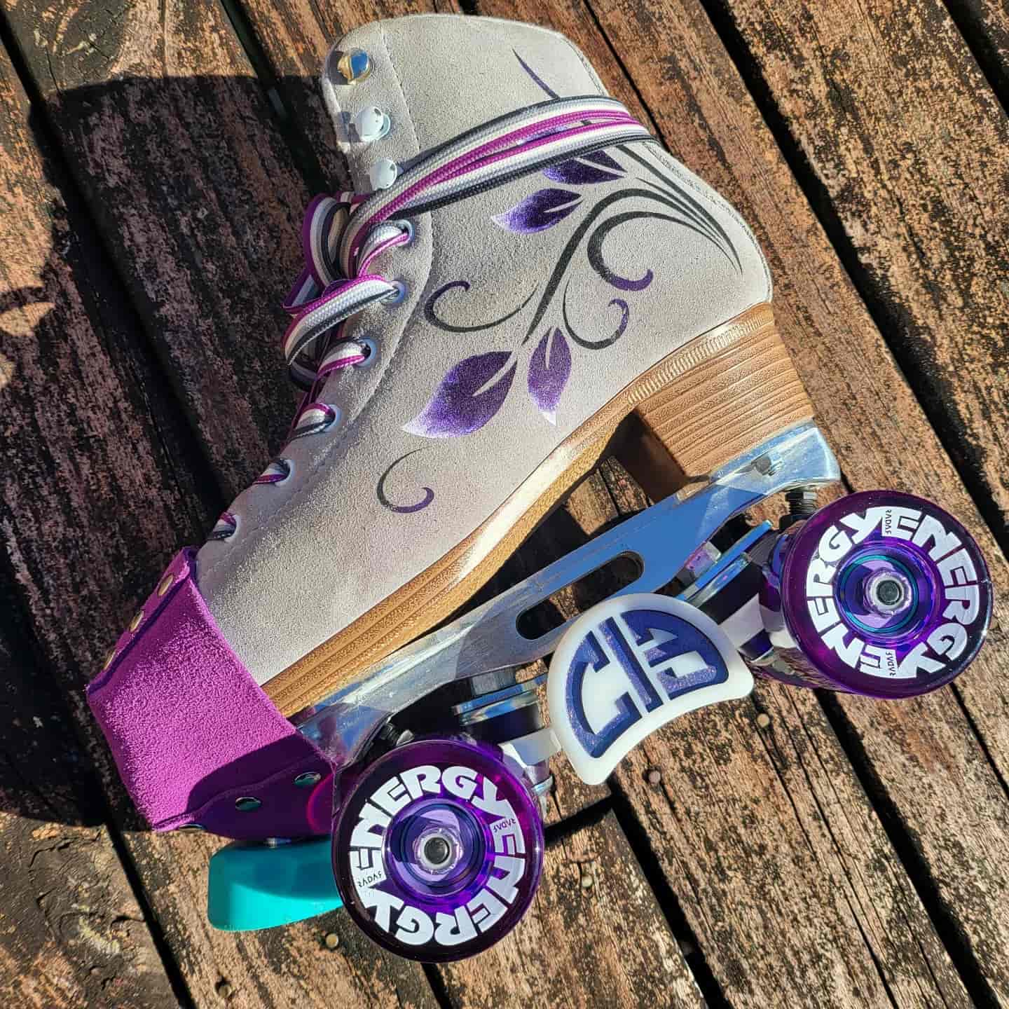 A grey suede rollerskate with purple wheels, teal toe stop, purple toe cap, and ace flag laces. The side of the skate is decorated in a floral leafy design in black, white and purple.