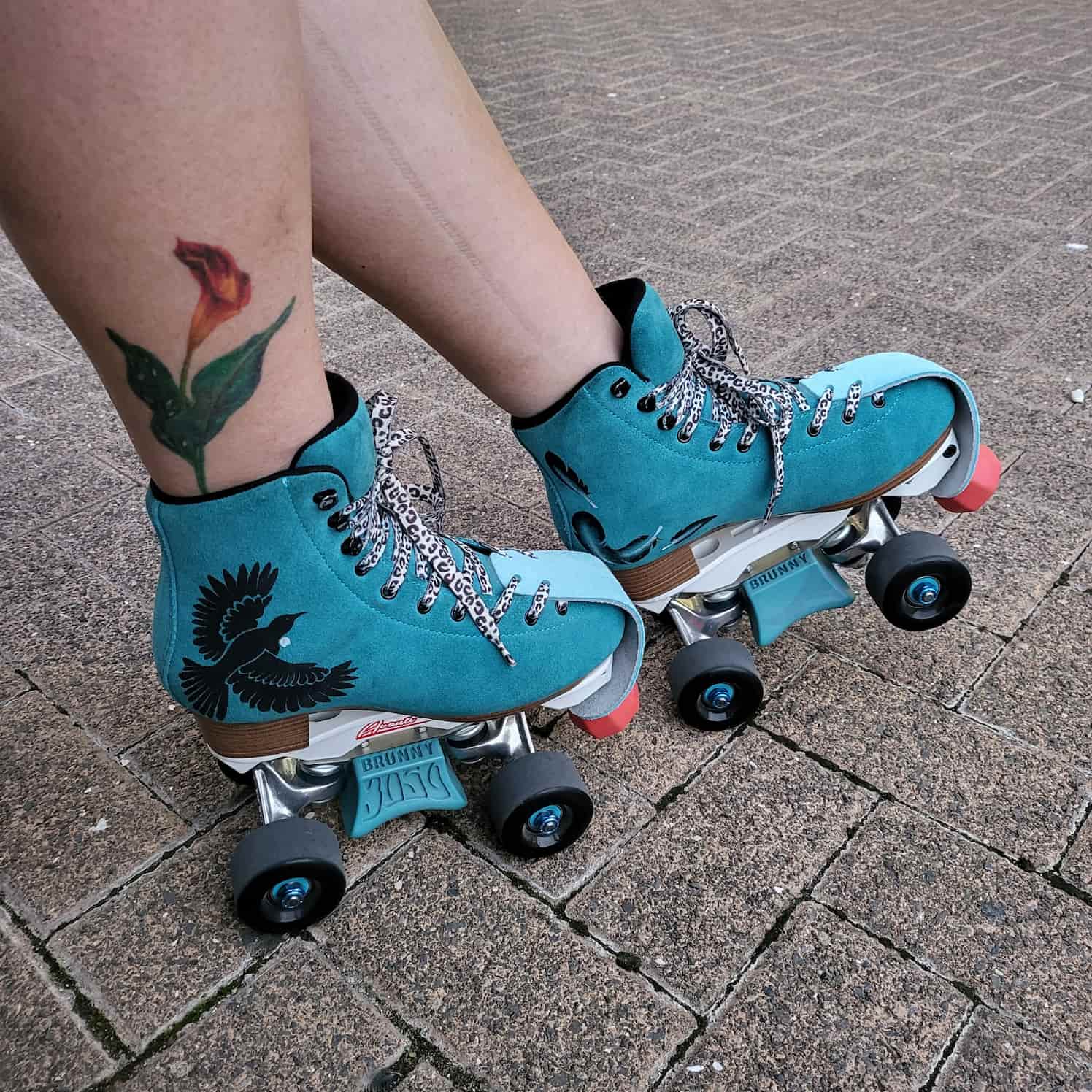 Teal blue suede rollerskates with black wheels, slide blocks, red toe stops, light blue toe caps, and leopard print laces. The side of one skate has a black tui design, the tui is in flight, it has the white throat decoration. The side of the other skate has three feathers in black and white.
