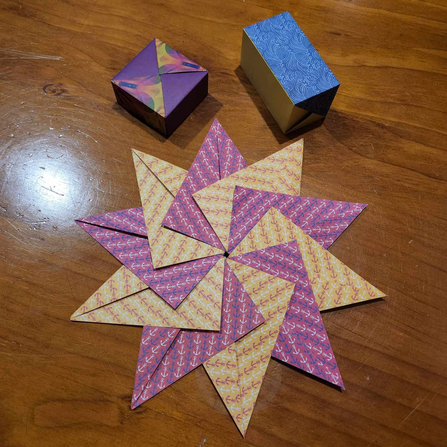 Modular origami star in anchor-patterned yellow and pink paper (alternating). It's lying flat on a wooden surface and there are some origami boxes in the shot as well.