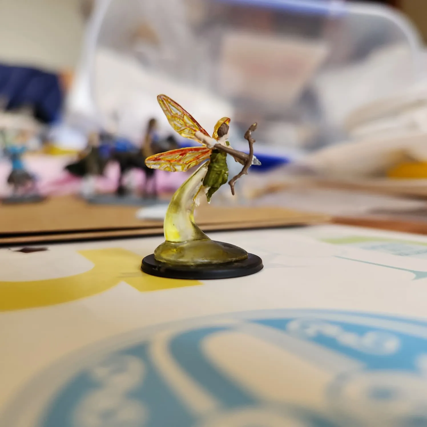 A tiny flying fairy figurine. The base is of transparent yellow, suspending the fairy. Her wings are transparent yellow with red veins. She has pale skin, brown hair, and green clothes. She is holding a bow with an arrow cocked in it.