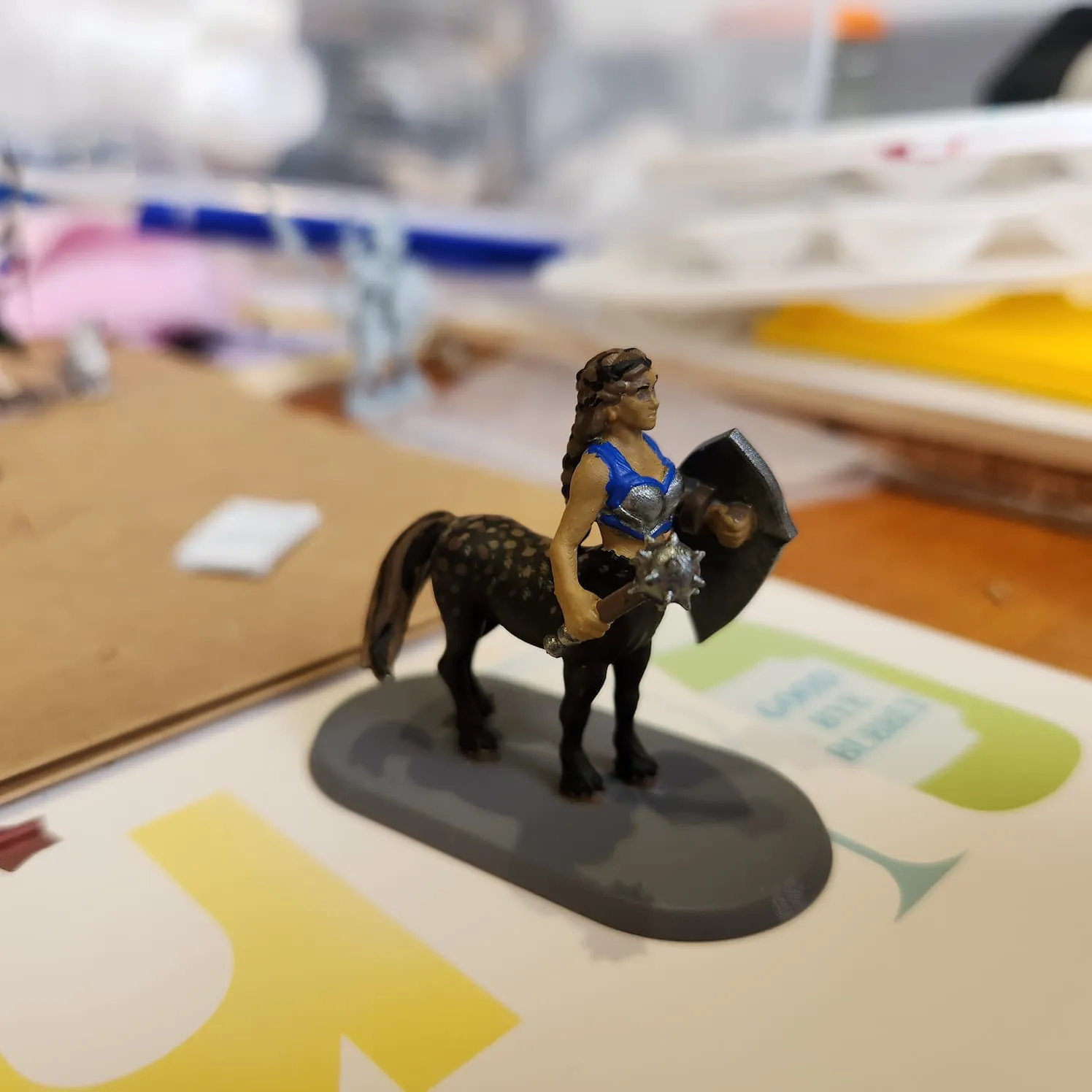 A female centaur standing steady on all fours. She has a dark horse body with lighter brown speckles on the back and streaked tail. Her hair matches the tail. Her human skin is mid-brown and she is wearing a blue and silver breastplate. In one hand she holds a shield, in the other a mace.