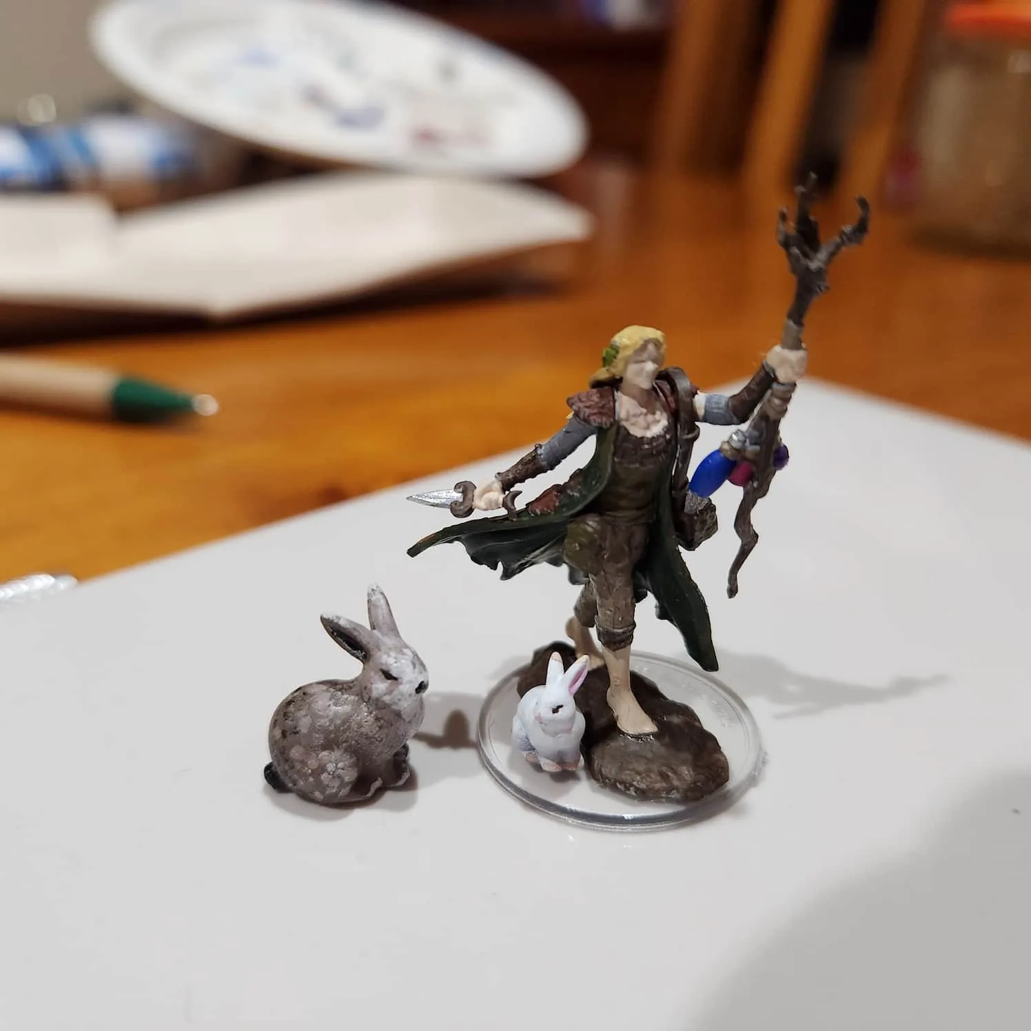 Minitature of a female human druid, barefoot and stepping forward as if moving. She is holding a wooden branch as a staff that has some items attached to it in her leading hand, and a dagger in her trailing hand. Her clothes are greens and browns. Her hair is blonde. At her feet are two rabbits - a white one, and a larger speckled brown one.