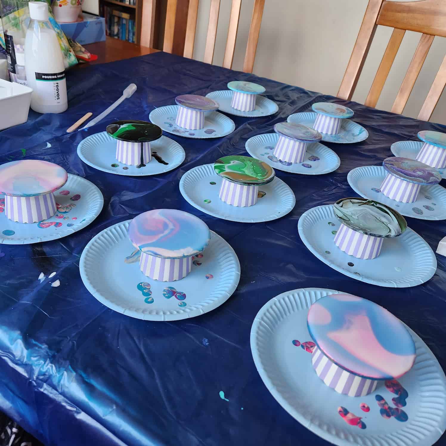 Wide shot of various coasters with acrylic paint poured on them, each standing propped up on a paper cylinder on a paper plate. The surface they are on is covered with a blue plastic cloth.