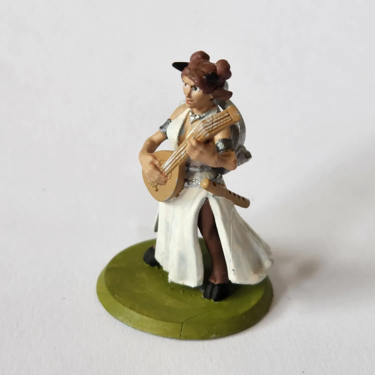 Female satyr bard miniature. She has light skin, medium brown hair in buns, and darker brown satyr legs with black hooves. She is playing a gitern, has a flute attached to her belt, and a lyre on her back. She has a white dress and silver armbands and belt.