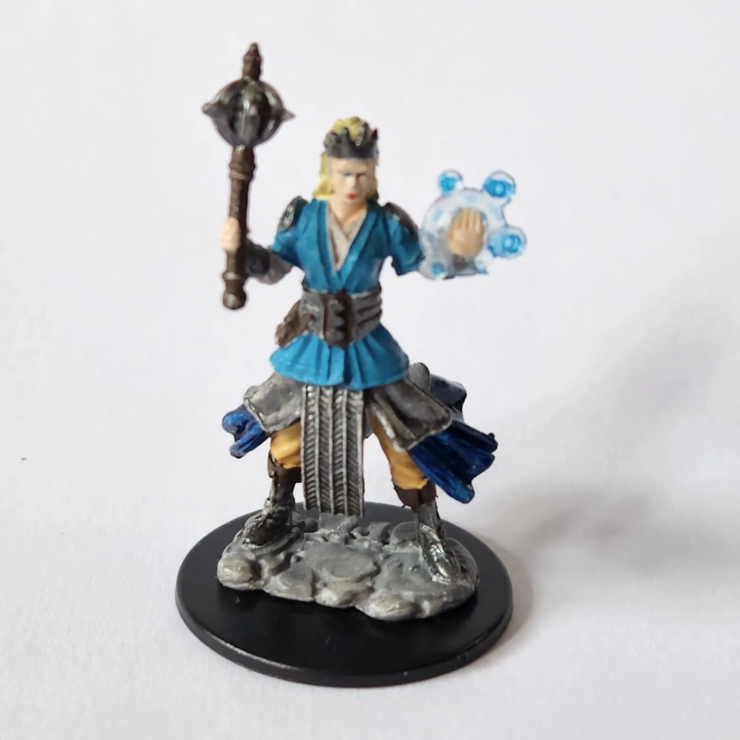 Miniature of a female human cleric. She has pale skin and yellow hair. She is standing solidly and holding a mace in one hand, and her other hand is palm out with a transparent shield or spell against it. She is wearing chainmail under an outer tunic in blue. Under the mail you can see a dark blue fabric layer, tan pants, and silver boots. She has a silver crown on.
