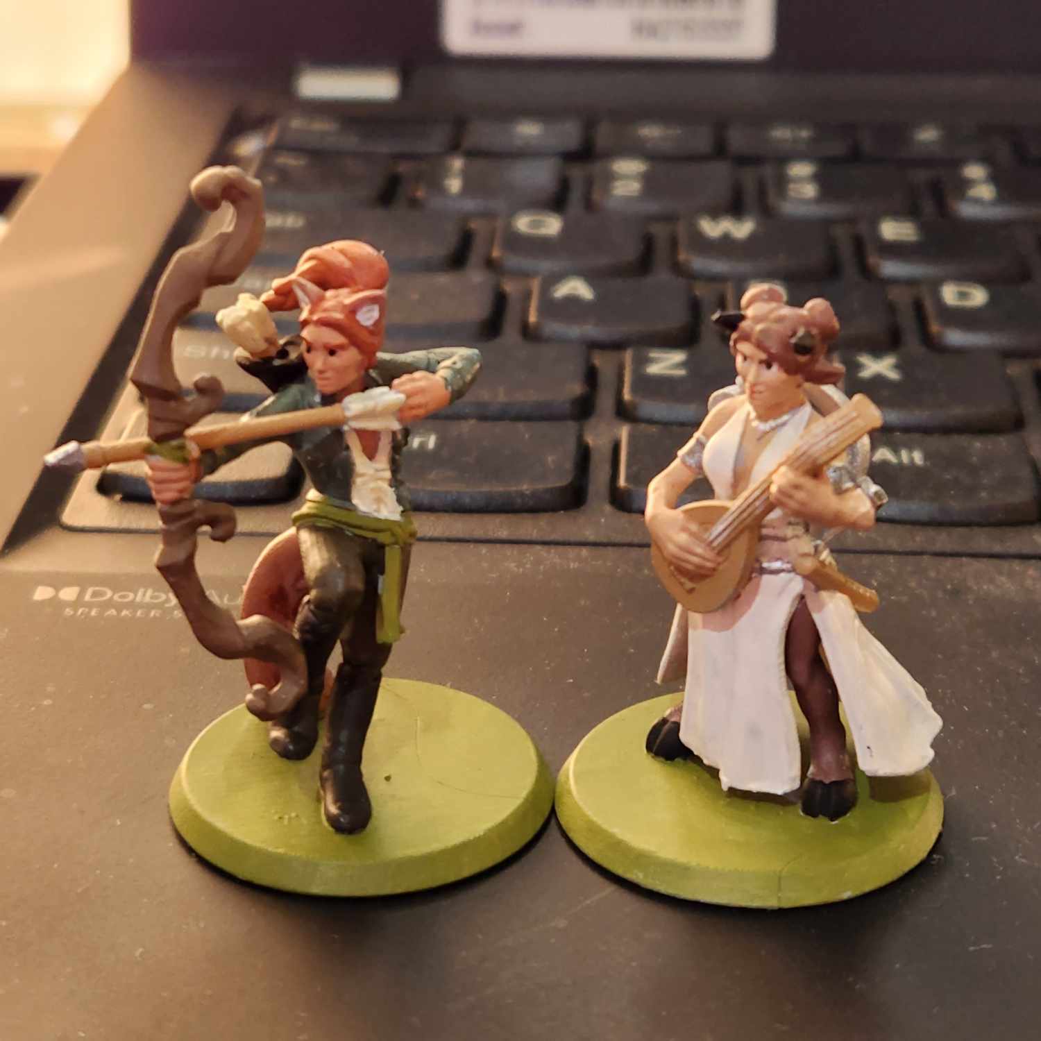Two miniatures stand on the surface of a black laptop, keyboard behind them. One is a human-fox archer, the other is a satyr bard.