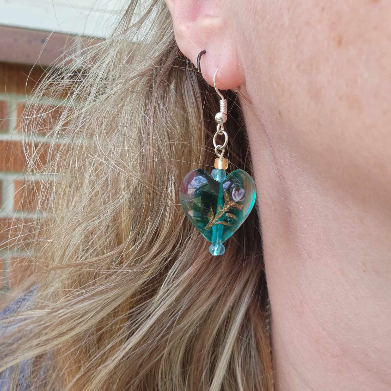 The glass earring is being worn. It hangs down from the earlobe. There are two small beads in gold and teal and then the large heart-shaped glass bead in teal, with a gold floral design on it.