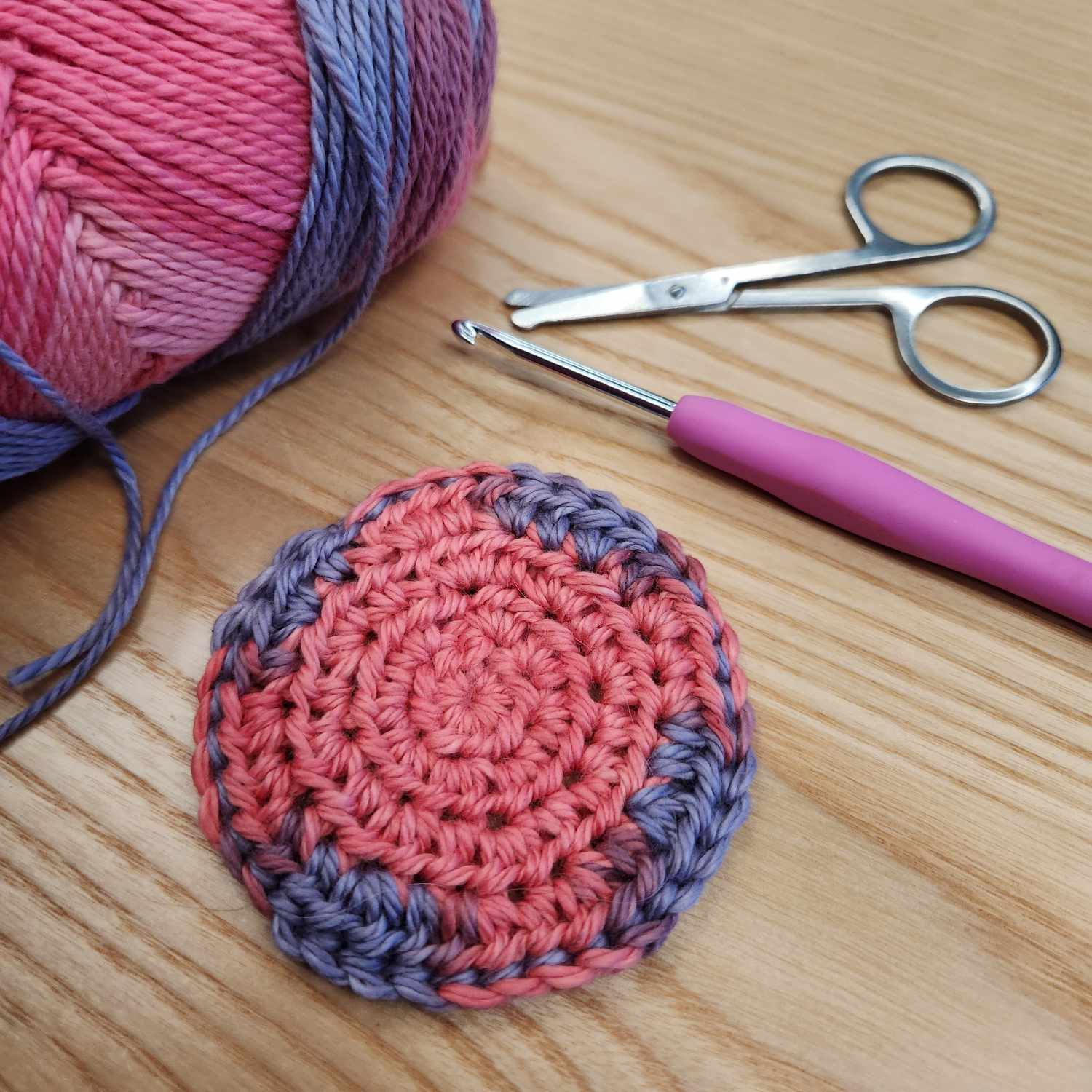 A crocheted circle in pink and purple yarn on a wood background. The pink-handled crochet hook, small scissors, and ball of yarn are also visible in the shot.