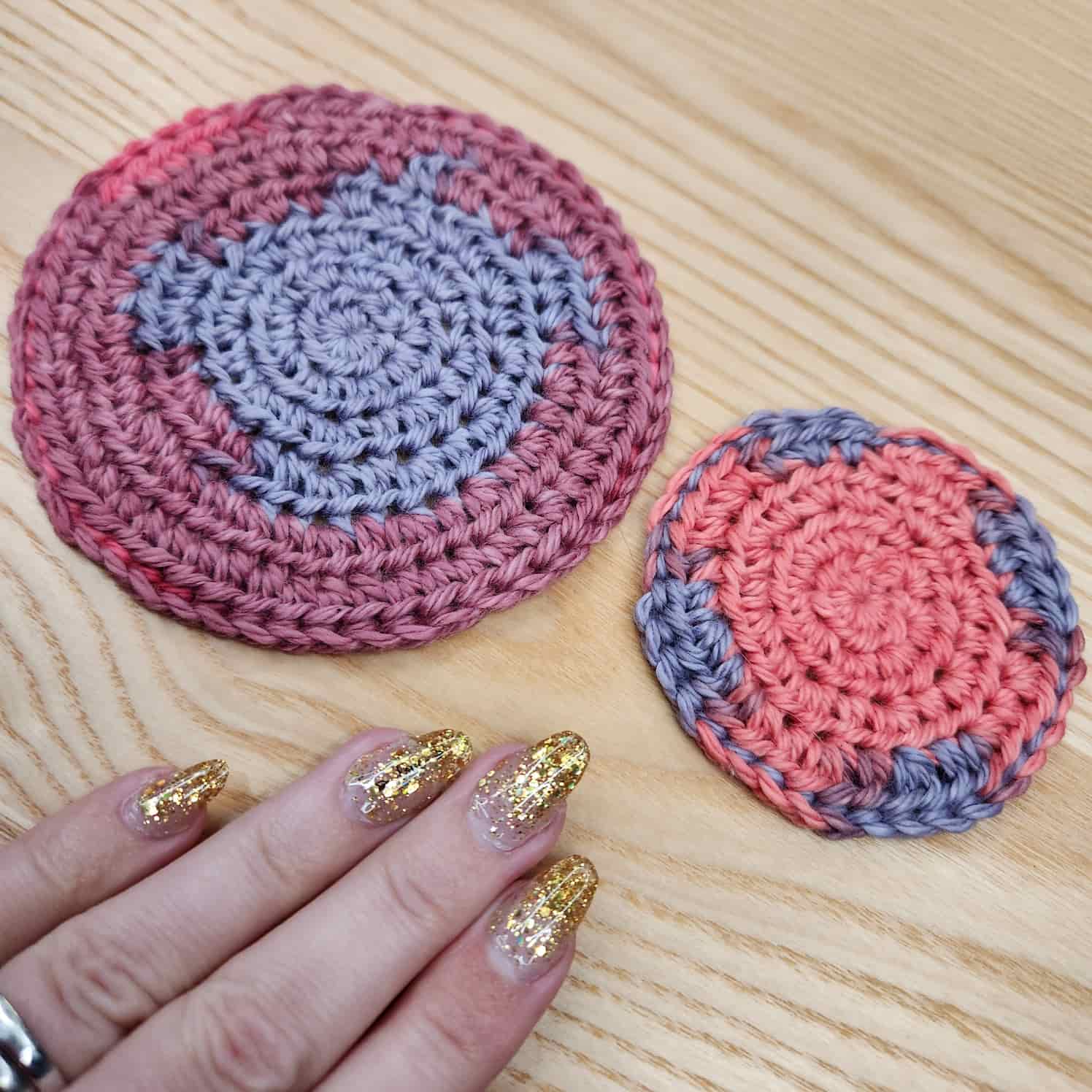 Two crocheted circles in pink and purple yarn on a wood background. A hand with gold nail polish is in the shot for scale, showing that the largest circle is about palm-sized, and the smallest is about three finger-widths.