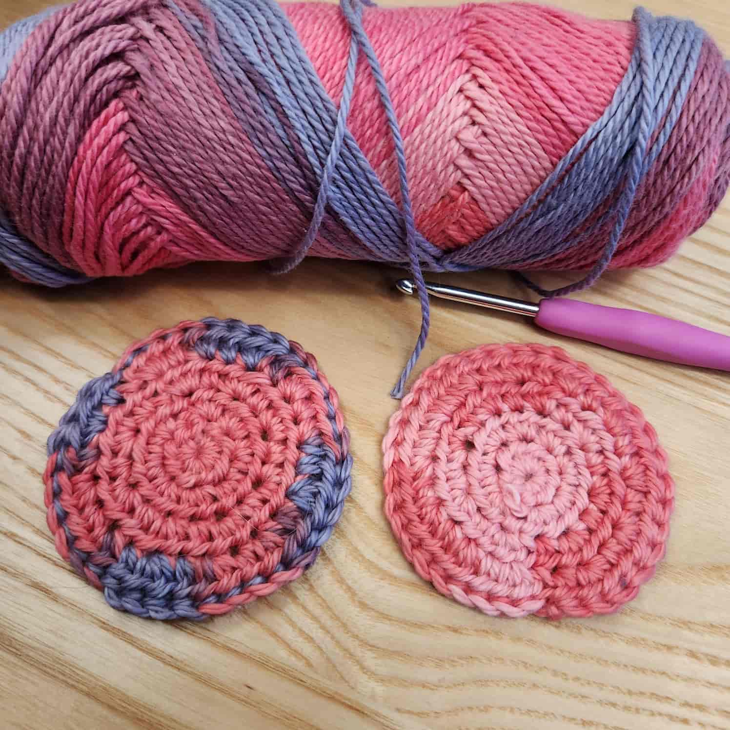Two crocheted circles in pink and purple yarn on a wood background. The pink-handled crochet hook and ball of yarn are also visible in the shot.