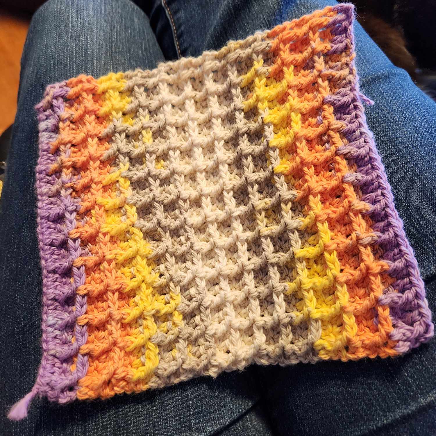 A waffle-stitch crocheted face cloth in colourful fabric of purple, orange, yellow, brown and off-white.
