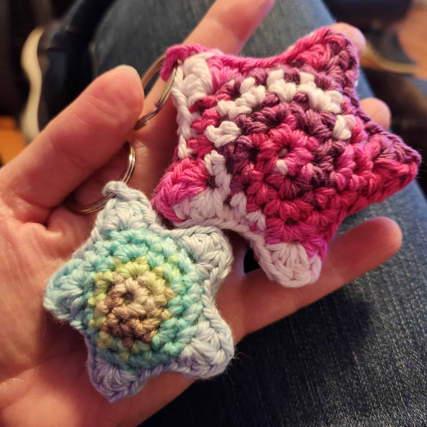 Two crochet stars on keyrings held in a hand. The blue, green and turqoise one is smaller, the pink and white one is about twice the size.
