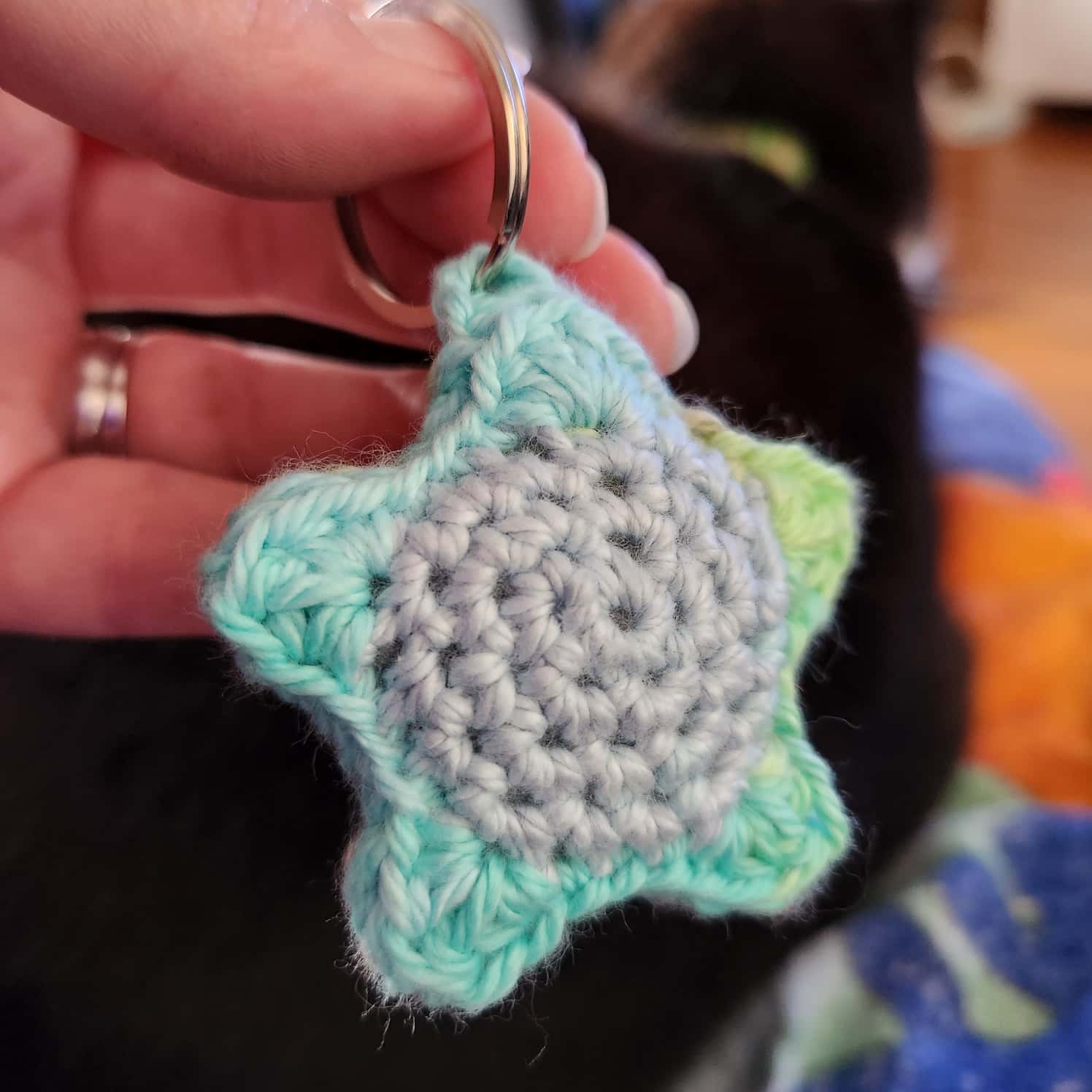 A green, blue and turquoise crochet star on a keyring held in a hand.