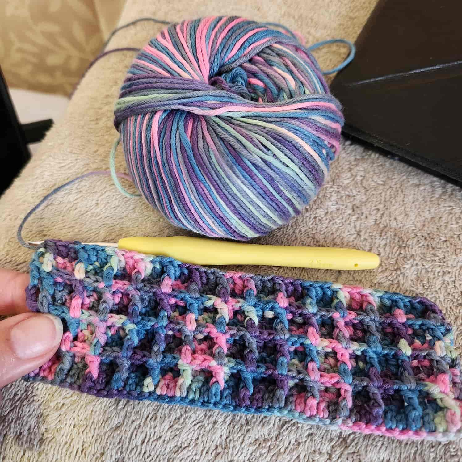 A work-in-progress white, teal, pink, purple and blue waffle stitch face cloth. Only a few rows are done, the yellow-handled crochet hook is still through a stitch loop. The ball of yarn is in shot.