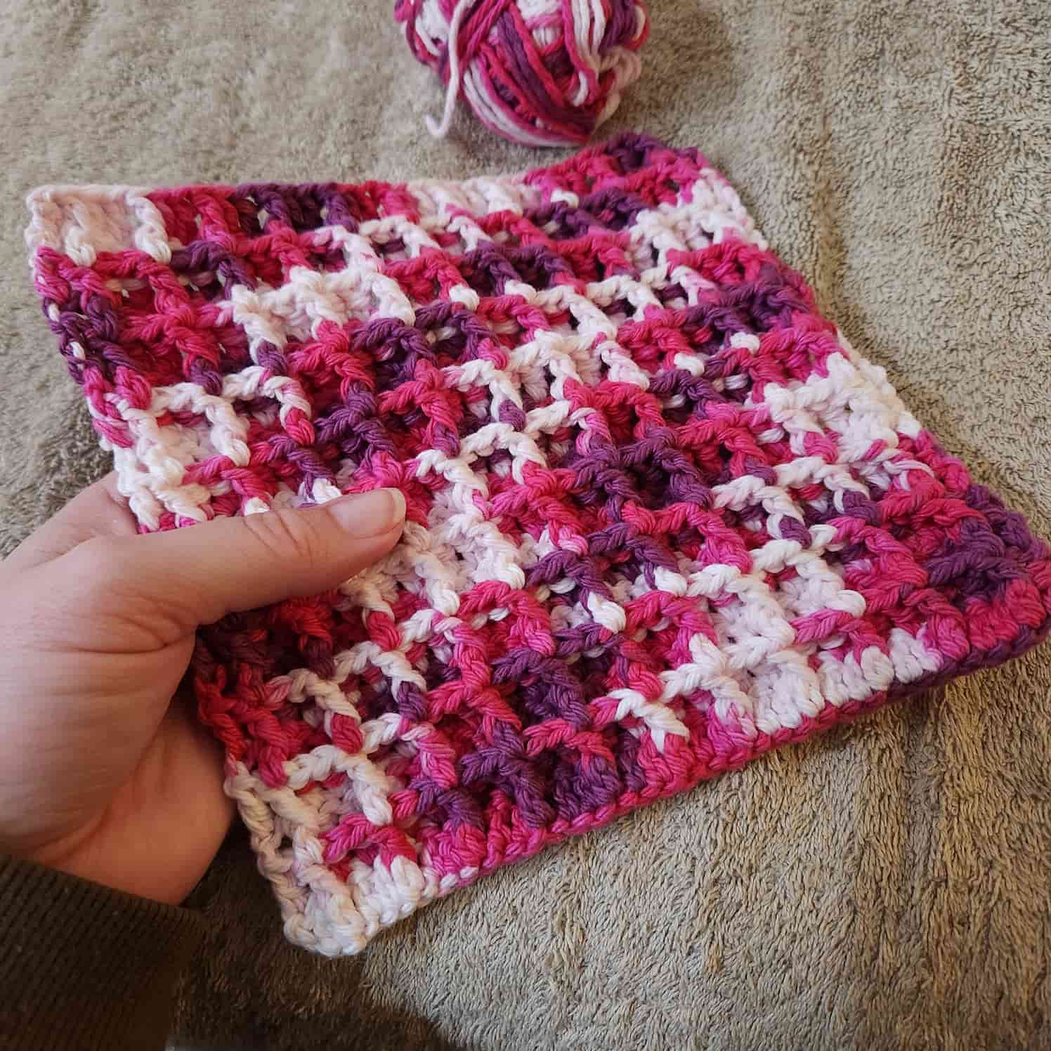 A completed pink, white and purple waffle stitch face cloth, held in a hand. The remaining ball of yarn is half in frame at the top.