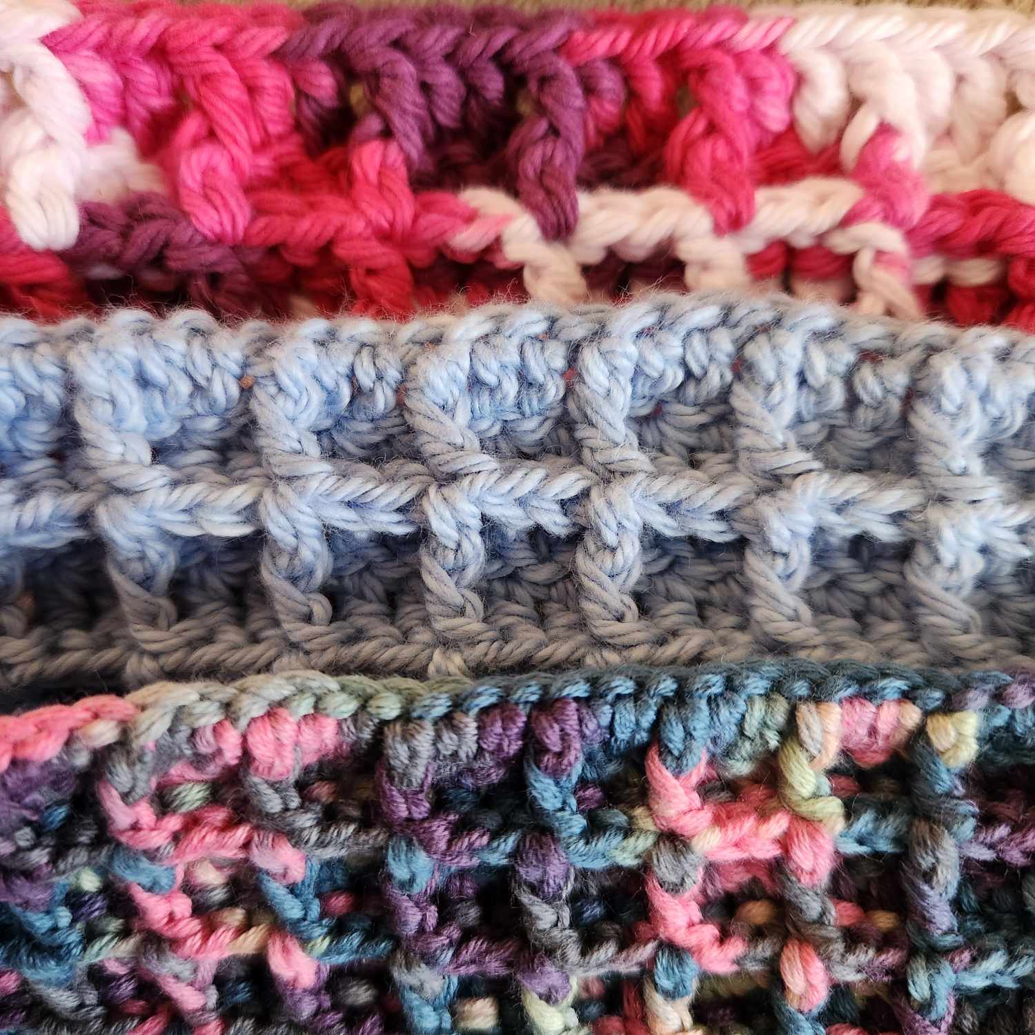 Three crochet waffle face cloths stacked. One is pink and white, one blue, the other a mixture of colours from white to pink to blue to purple.