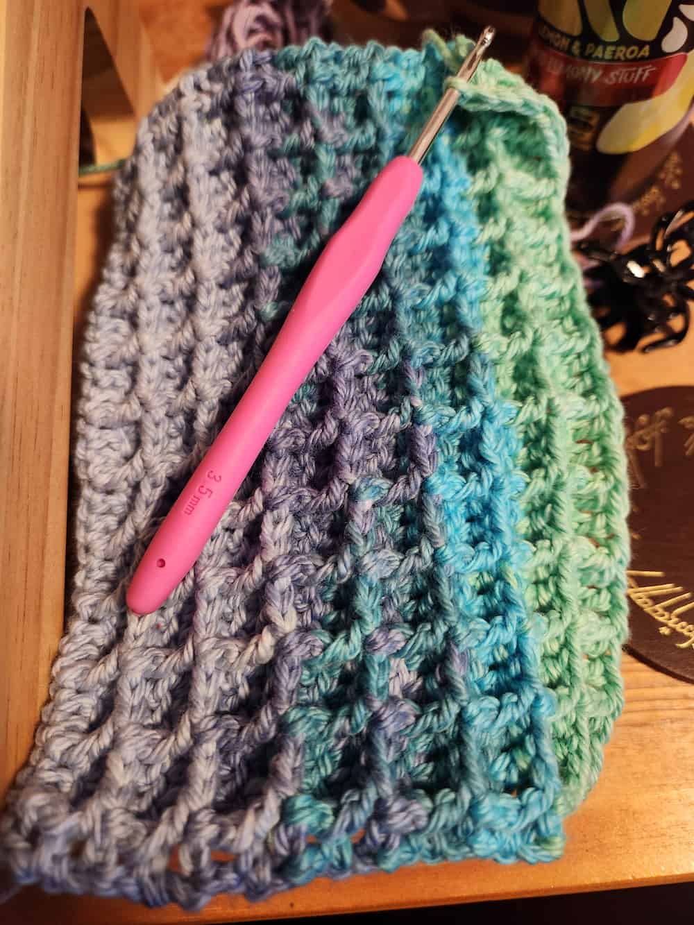 Work-in-progress purple, blue and green waffle stitch crochet face cloth. The pink-handled crochet hook is still through a stitch loop.
