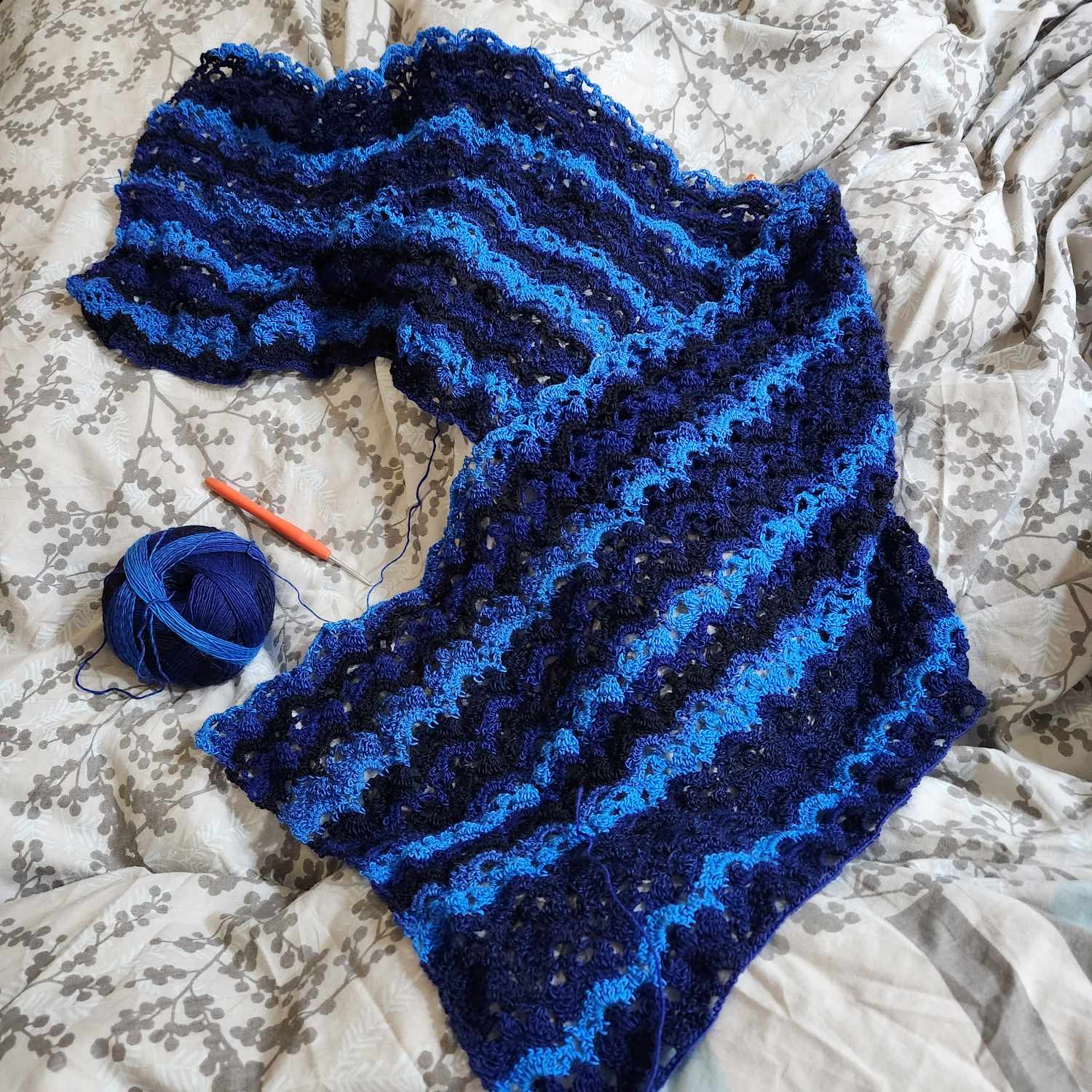 A work-in-progress crochet project of a rectangular shawl in a lacey pattern. The yarn gives the shawl light blue, dark blue and black stripes. The orange-handled crochet hook and ball of yarn are in the shot. The shawl is nearing completion at this point.