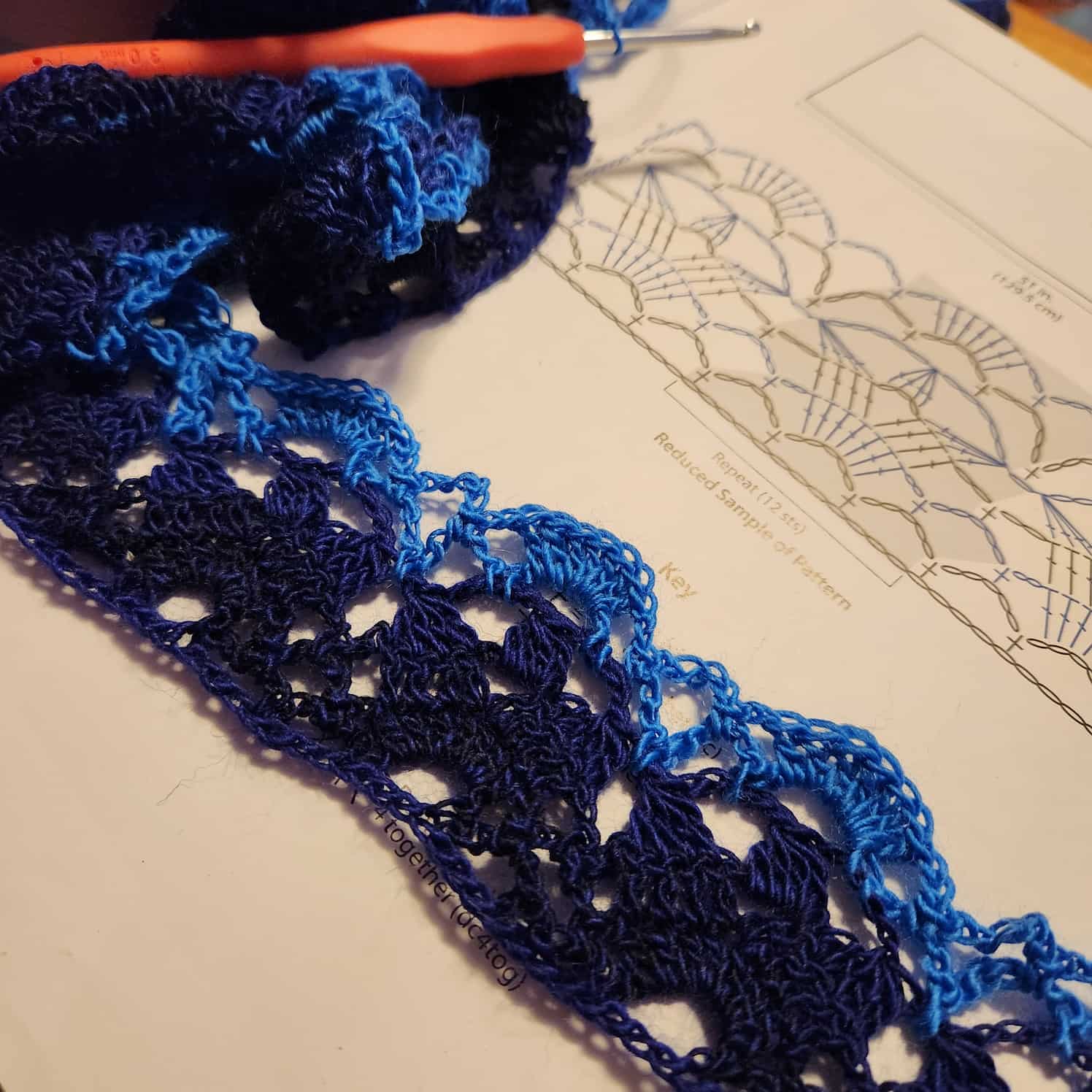 The very start of the blue lacy crochet shawl. The first few rows are done, the orange-handled crochet hook can be seen in the upper left corner, and the pattern is shown.