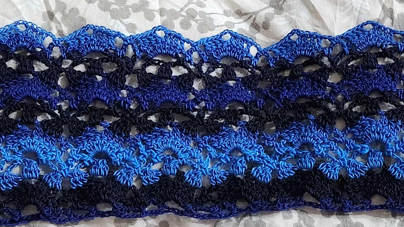 Progress shot of the shawl with some more rows done. The image shows off the nice lace pattern and the light blue, dark blue, and black stripes of the yarn.