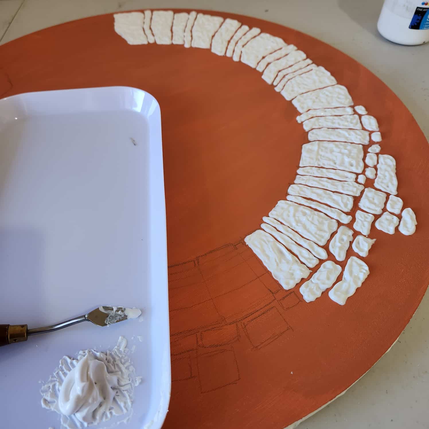Round stretched canvas painted orange. A design is sketched on in pencil, and some of it has been filled with modelling paste. A plastic palette with modelling paste and palette knife sits on the canvas.