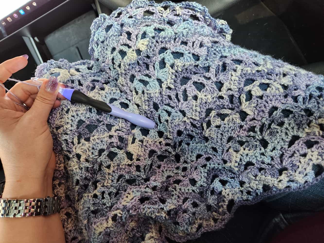 Work-in-progress shot of the shawl. The yarn changes from off-white to light blue to light and medium purple. The shawl is draped over legs, and a hand holds the lighted crochet hook and yarn off to the left.