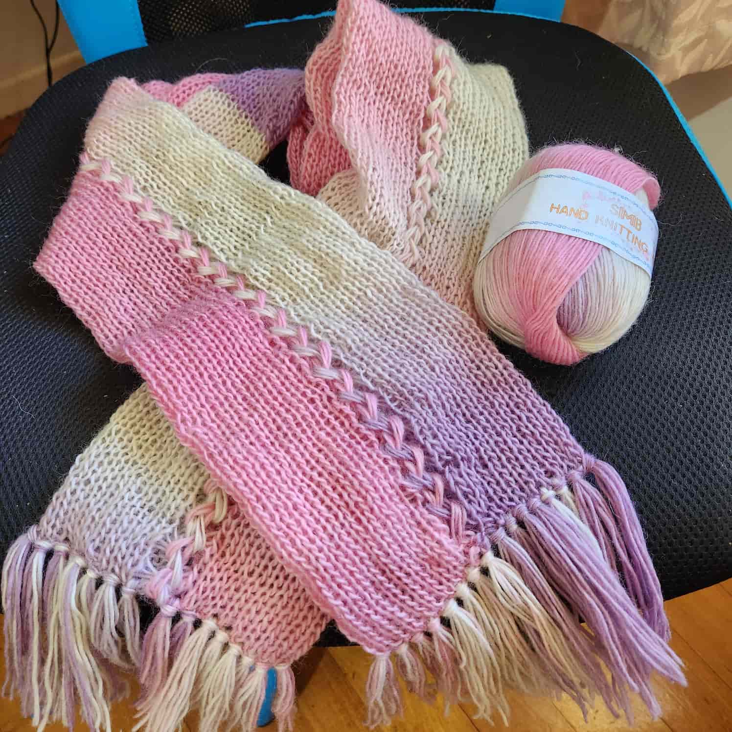 Scarf made from pink, purple and cream acrylic yarn. It has a braided design down the middle, and tassled ends.