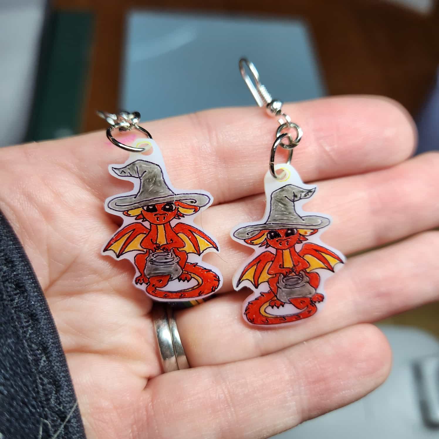 Close-up of a pair of earrings on white shrink plastic, a red dragon with yellow wings is wearing a black witches hat and holding a black cauldron.