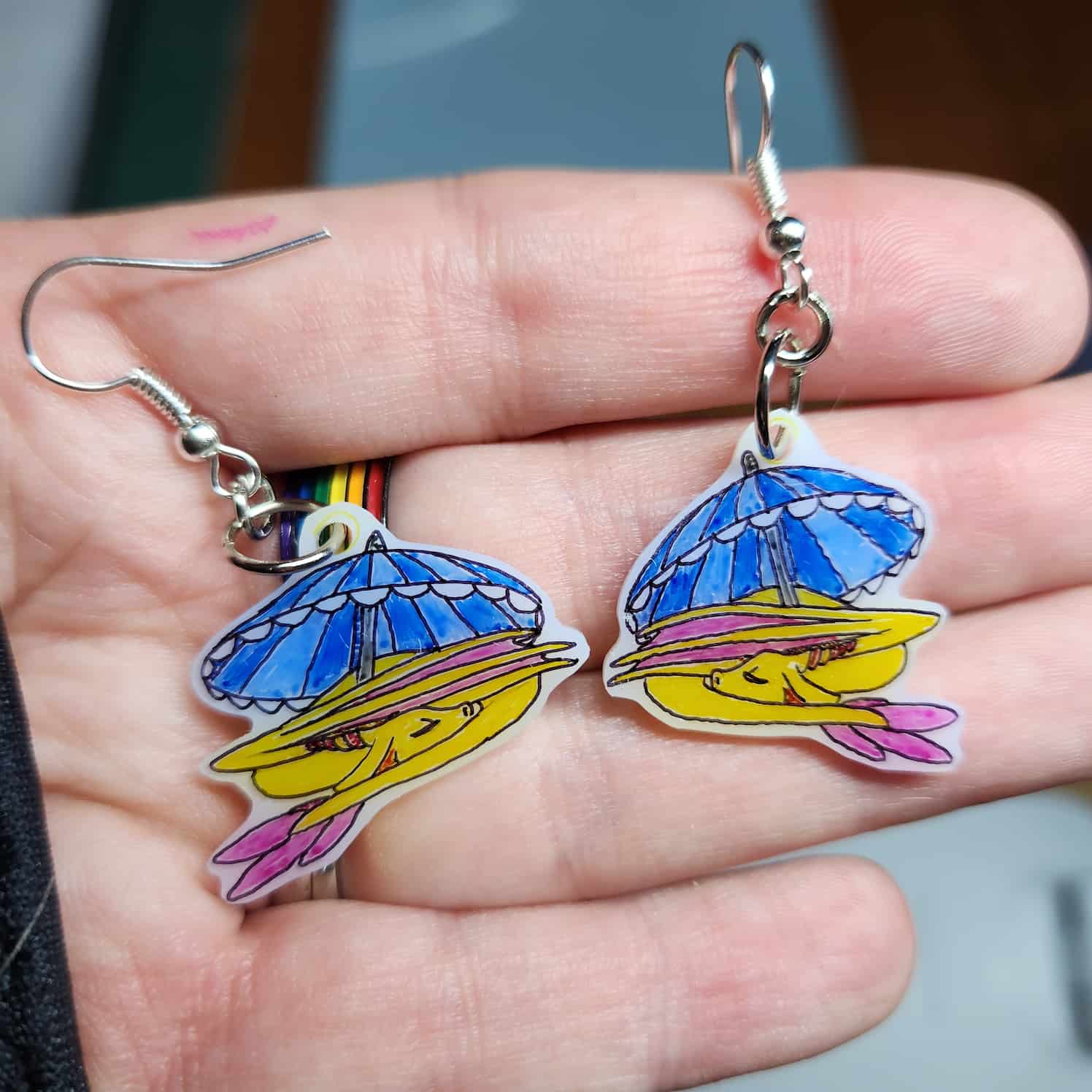 Close-up of a pair of earrings on white shrink plastic, a yellow dragon with pink wings and tail is curled up asleep under a blue striped beach umbrella.ß