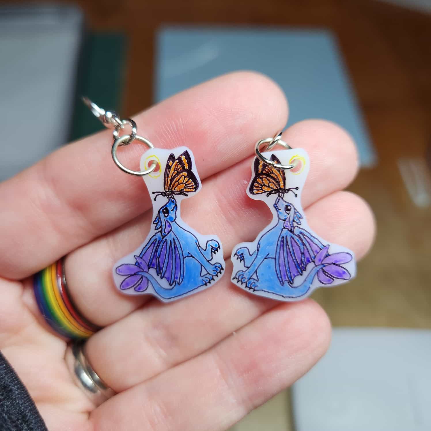 Close-up of a pair of earrings on white shrink plastic, a blue dragon with purple wings and tail is looking up at a butterfly on its nose.