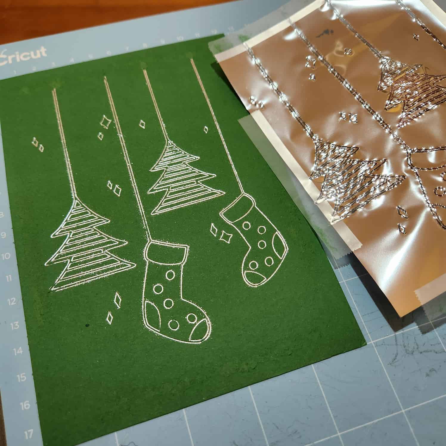 A green piece of card with a design in silver foil on it of Christmas tree and Christmas stocking ornaments hanging down from the top of the card. To the right is the discarded piece of foil.