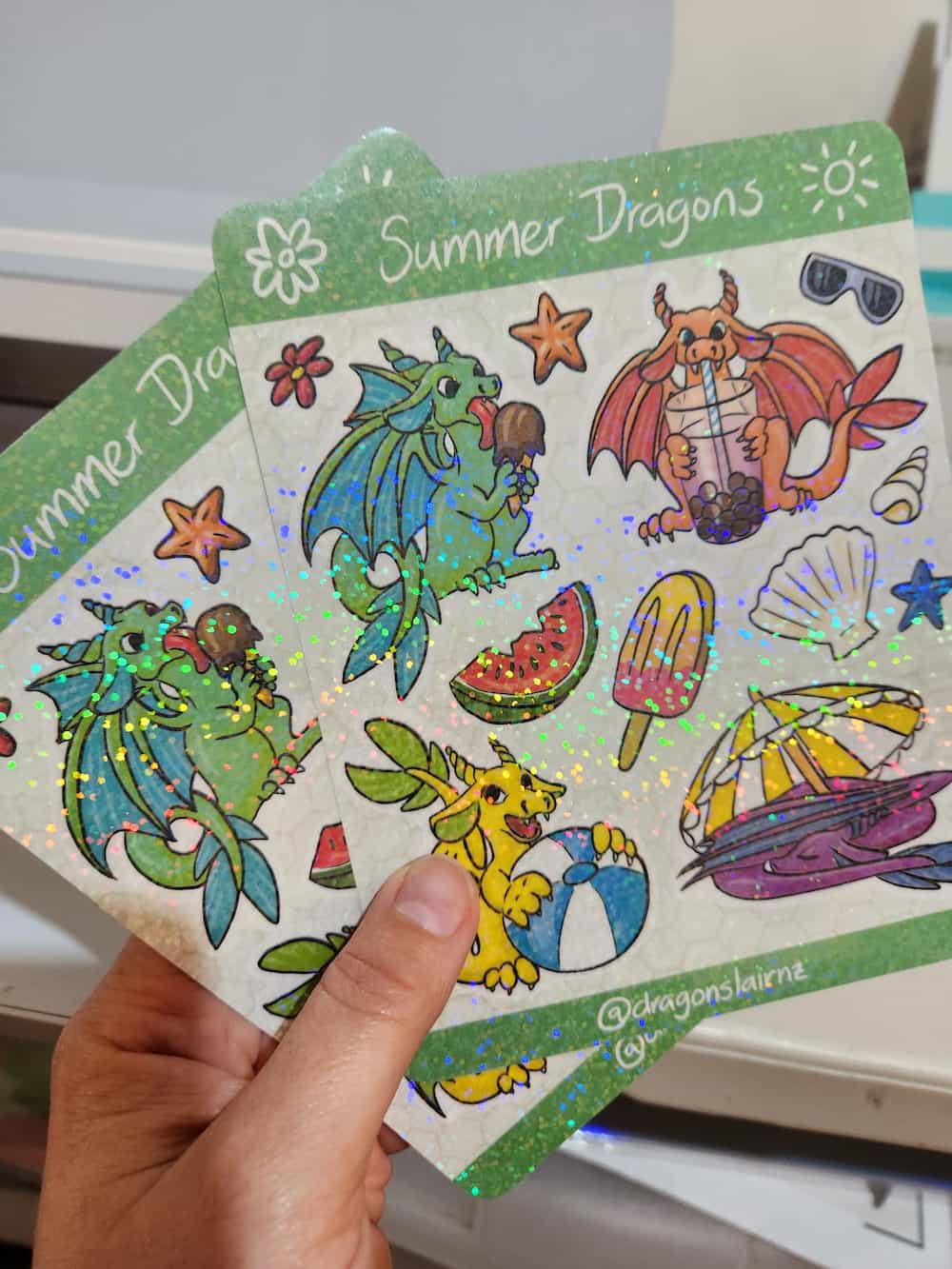 A hand is holding two sticker sheets titled "Summer Dragons" with dragons licking ice cream, drinking bubble tea, holding a beach ball, and curled up under a sun umbrella. Other summer-themed items are included like shells, popsicle, and watermelon.
