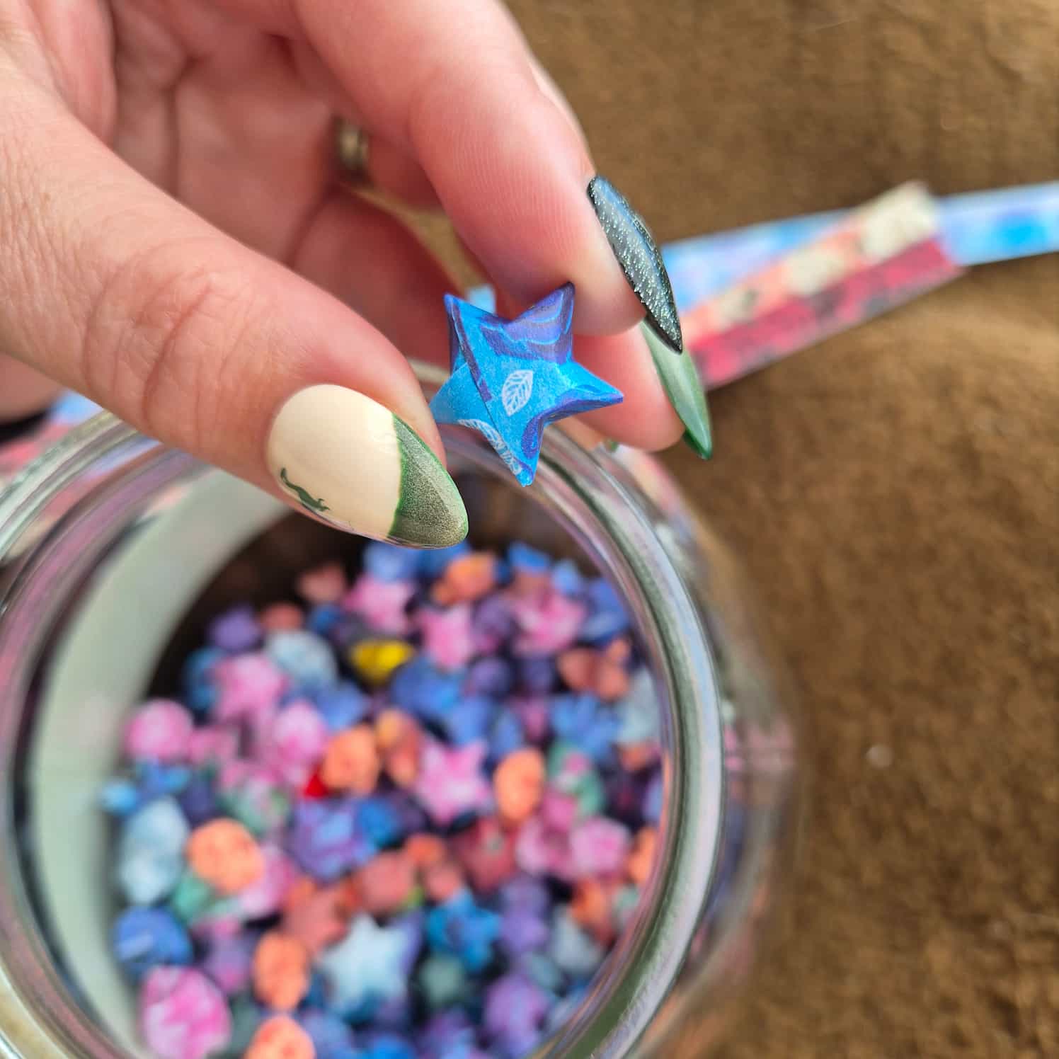 A hand holds a small origami star up to the camera. Unfocused in the background is a jar with many more folded stars, and some strips of paper ready to be folded.