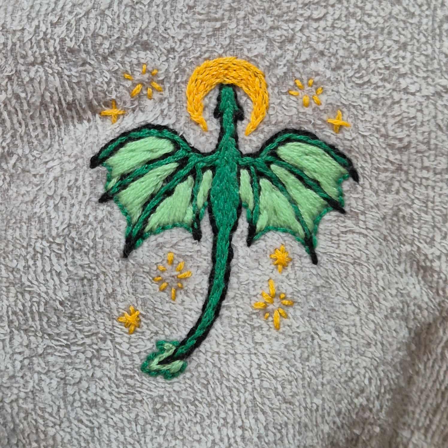 Close-up of a green dragon with yellow moon and star designs around it, embroidered onto a grey towel-material surface.