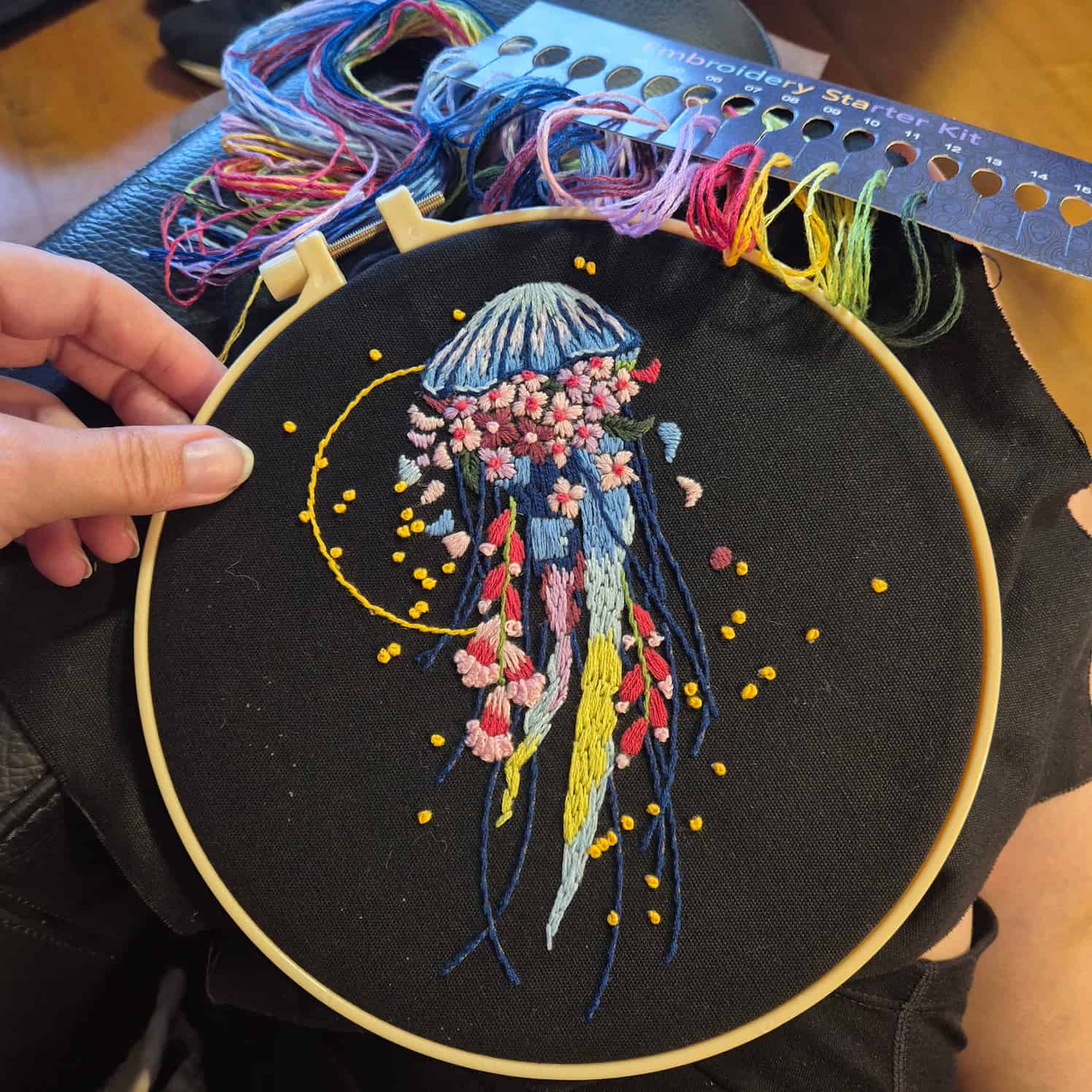 A hand holds an embroidery hoop with black fabric and the completed design of a colourful jellyfish with floral decorations. A card of colourful thread is included in frame.