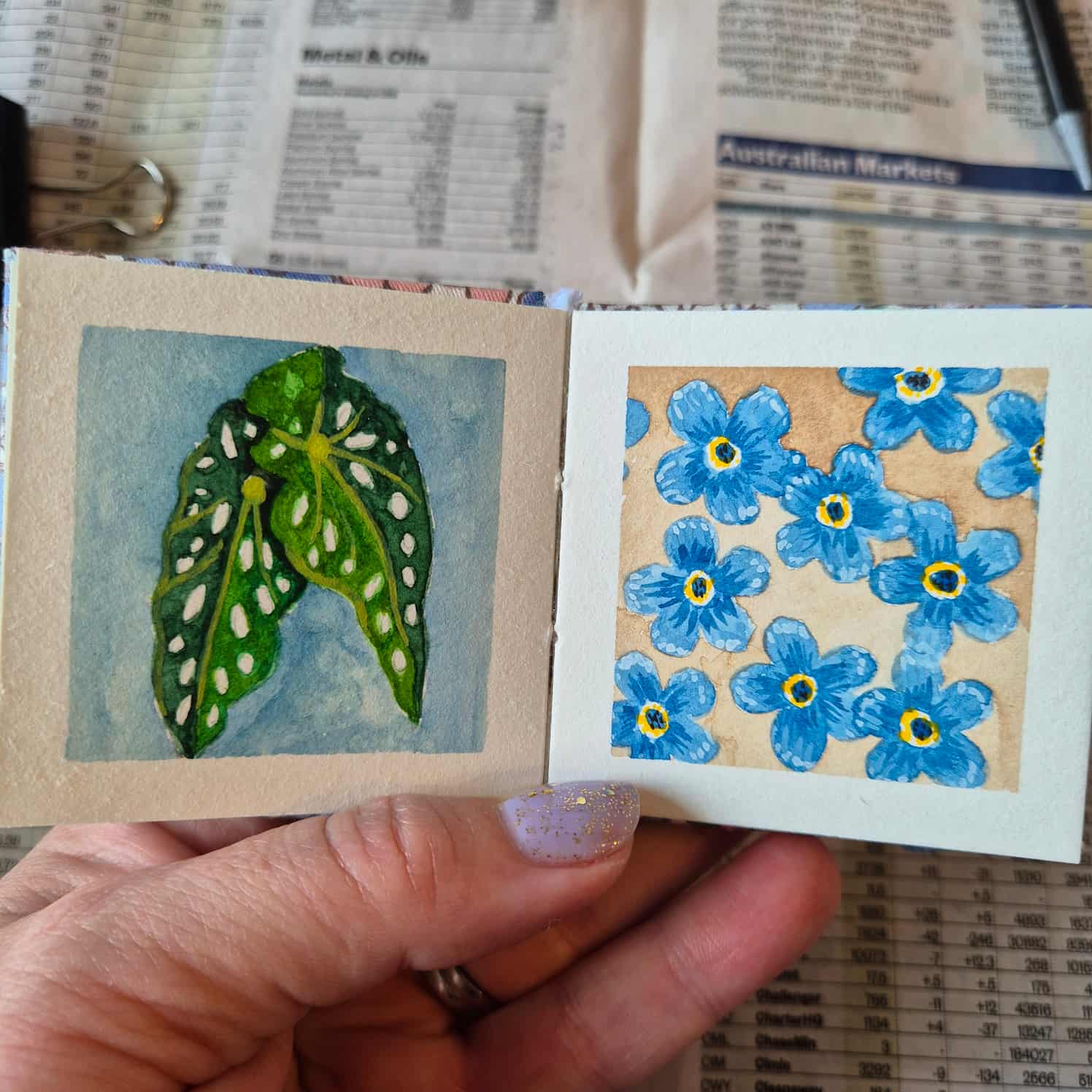 A small watercolour book open to two paintings - some green leaves on a blue background, and some blue flowers on a brown background.