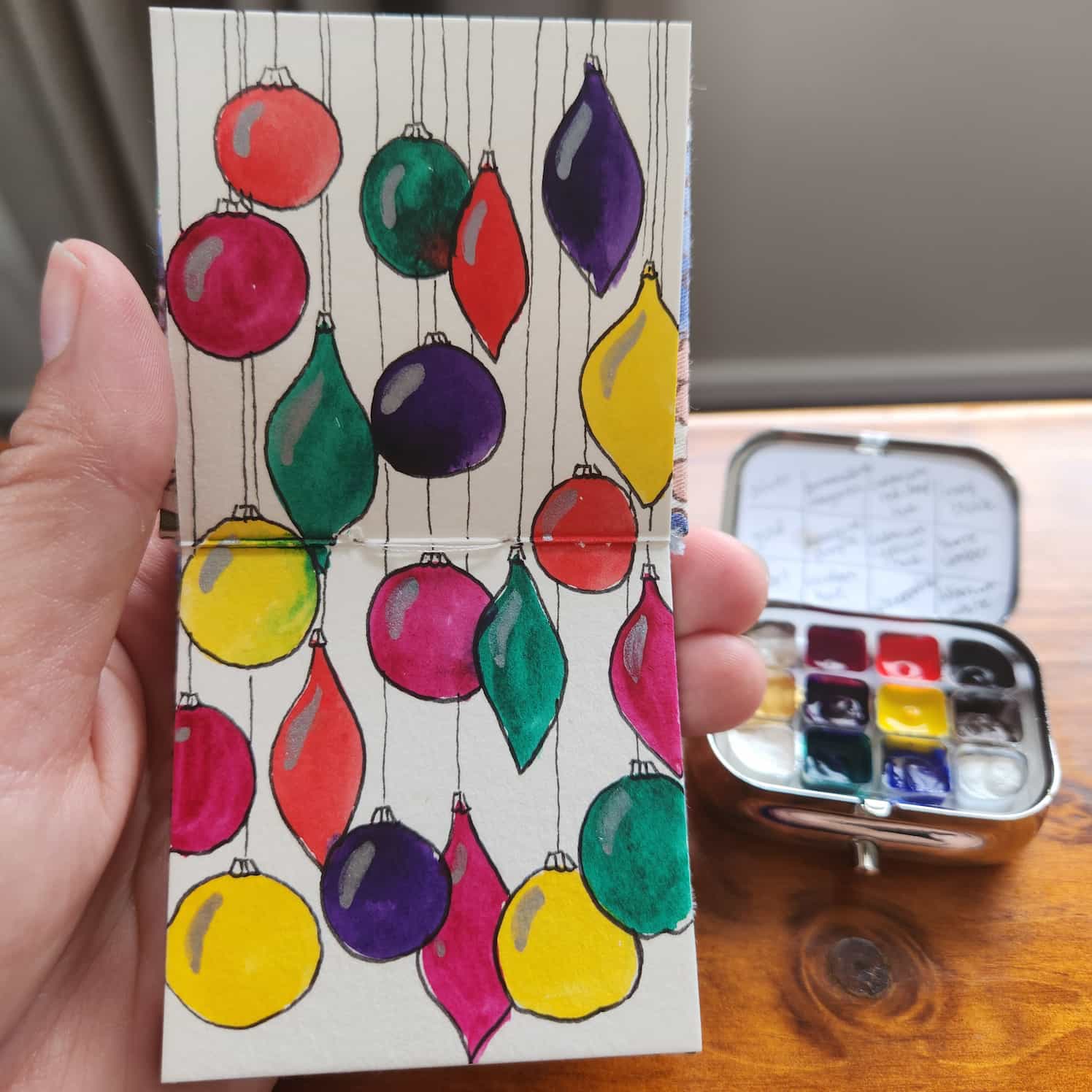 A small watercolour book open to a two-page spread of hanging colourful Christmas ornaments. They are overlapping in places, outlined in black, with silver highlights.