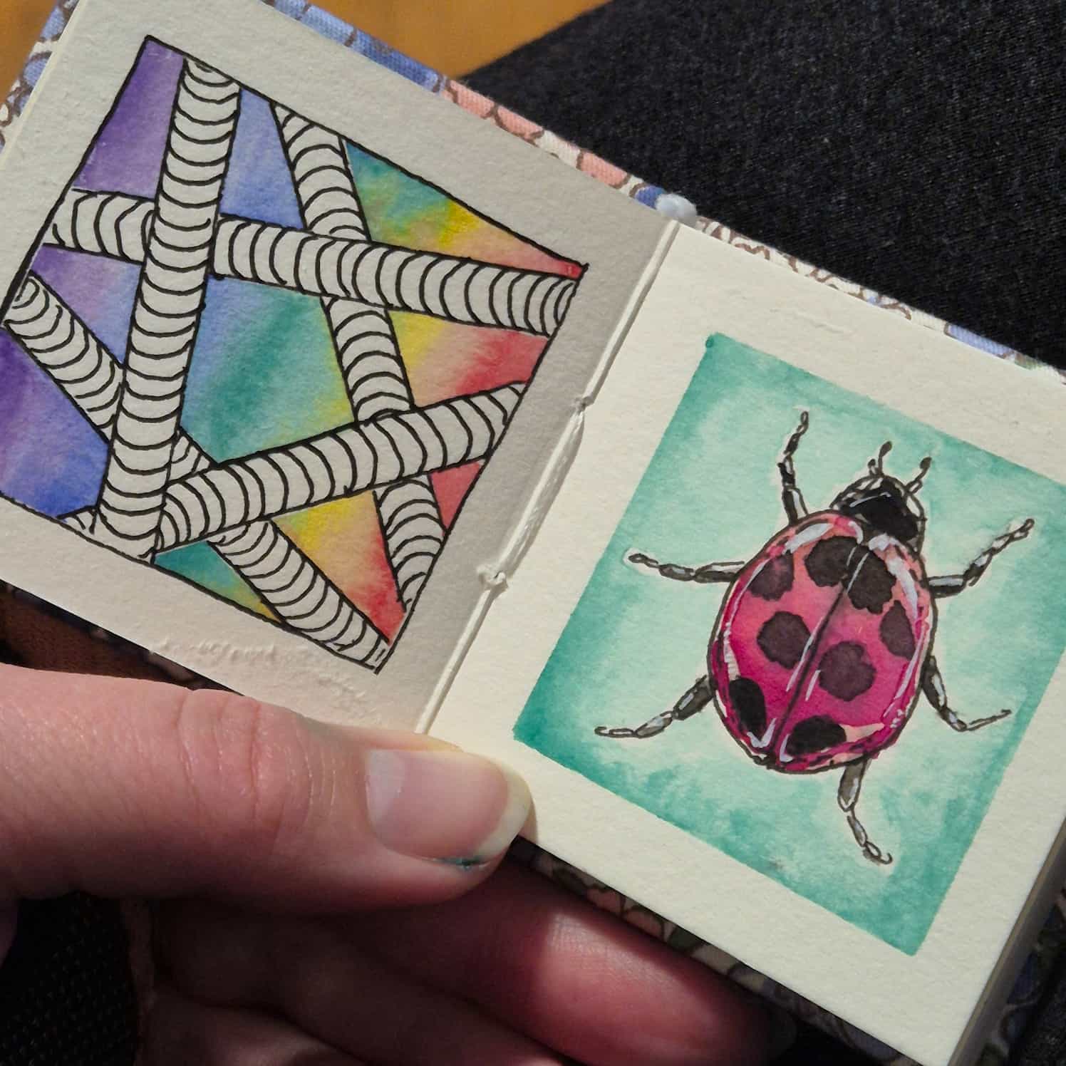 A small watercolour book open to two paintings - some black and white pipes with a rainbow background, and a shiny ladybird beetle on a green background.