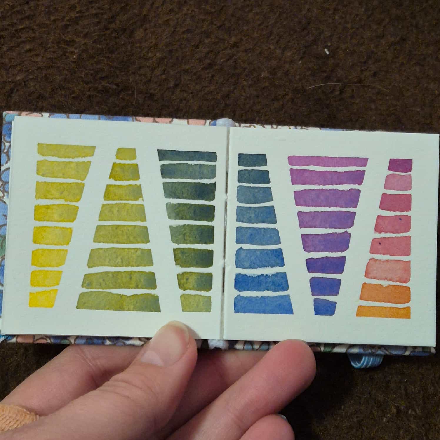A small watercolour book open to a two-page spread of lines going from a gradient of yellow to green to blue to purple to pink to orange.