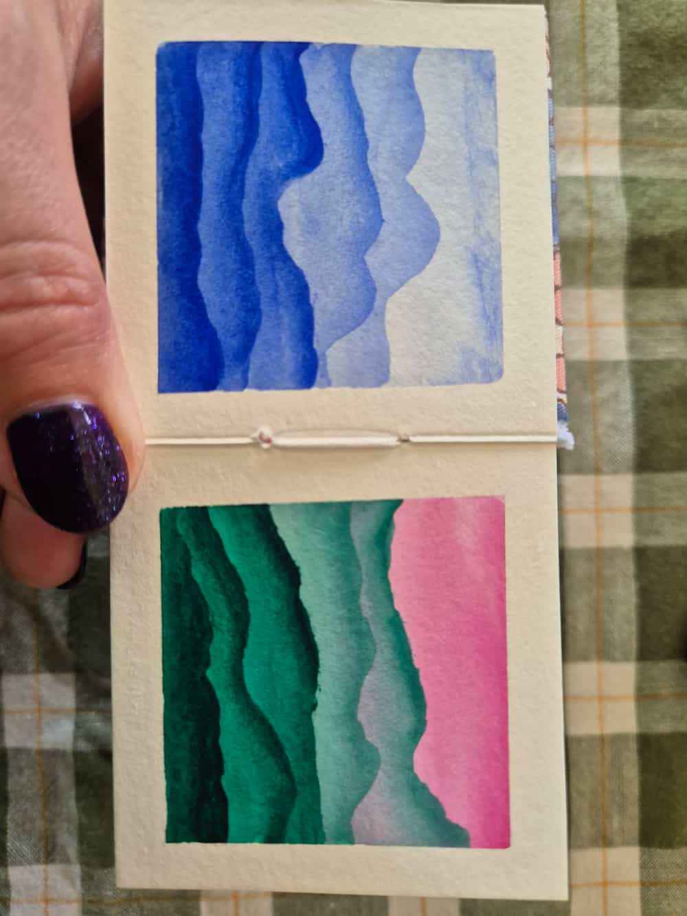 A small watercolour book open to two pages, both showing simple layered landscapes - one in all blue, the other in green and pink.