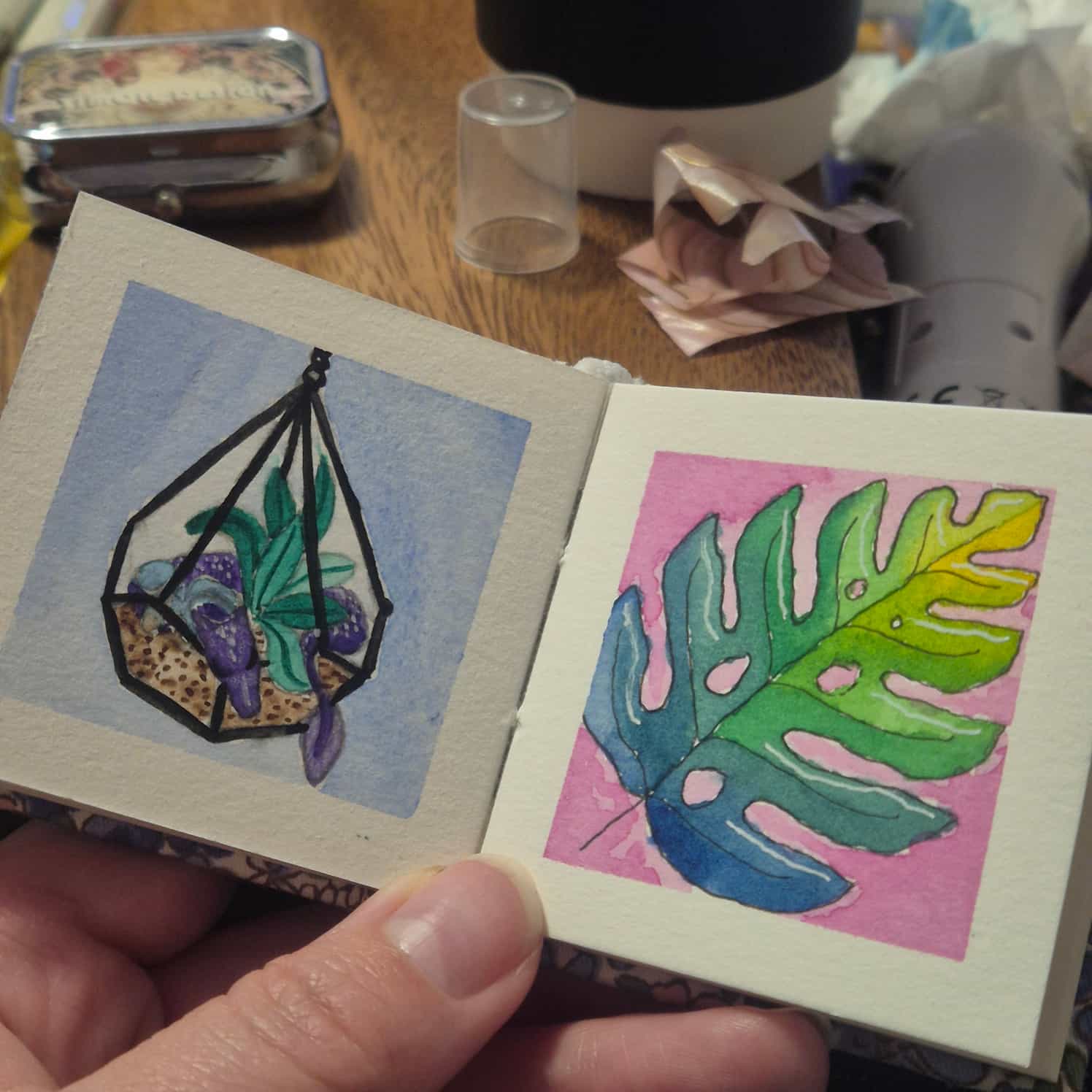 A small watercolour book open to two paintings - a small purple dragon curled up asleep in a terrarium, and a green-blue leaf on a pink background.