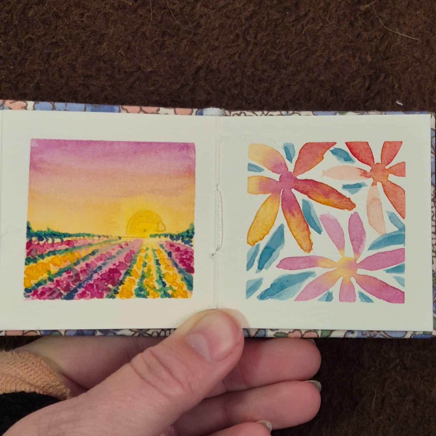 A small watercolour book open to two paintings - a field of rows of flowers in pink and yellow leading to a pink and yellow sunset sky, and some simple gradient flowers with leaves.