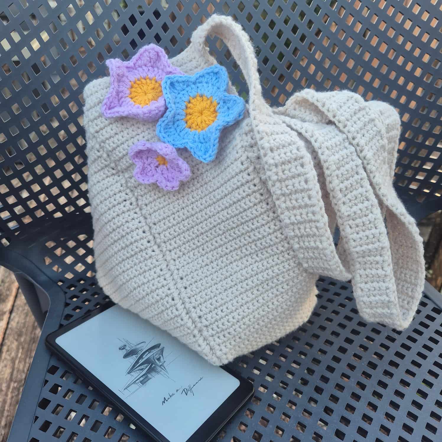 An off-white or cream crocheted handbag with pink and blue crocheted flowers attached near one strap sits on a grey chair. There is a small phone-like device on the chair next to it.