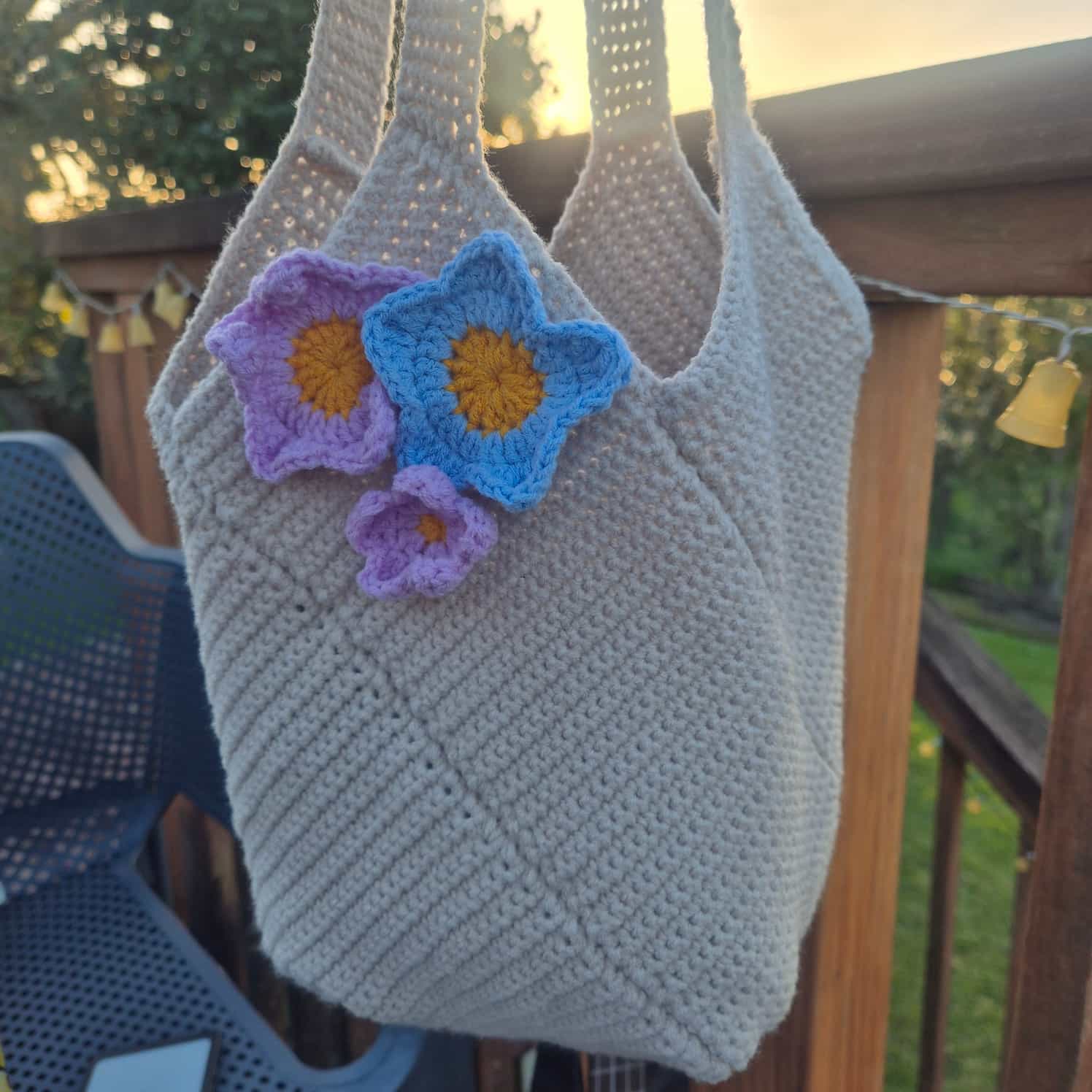 An off-white or cream crocheted handbag with pink and blue crocheted flowers attached near one strap is held up by the straps.