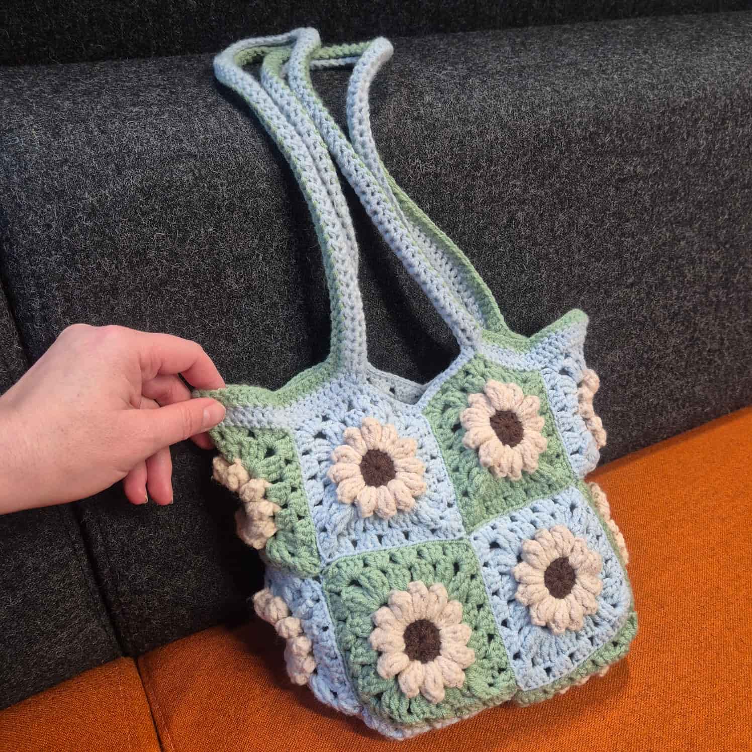 A handbag made of crocheted granny squares sits on a grey and orange background, a hand is included for scale. The squares are alternating light blue and green. They each contain a flower with cream petals and a brown center.